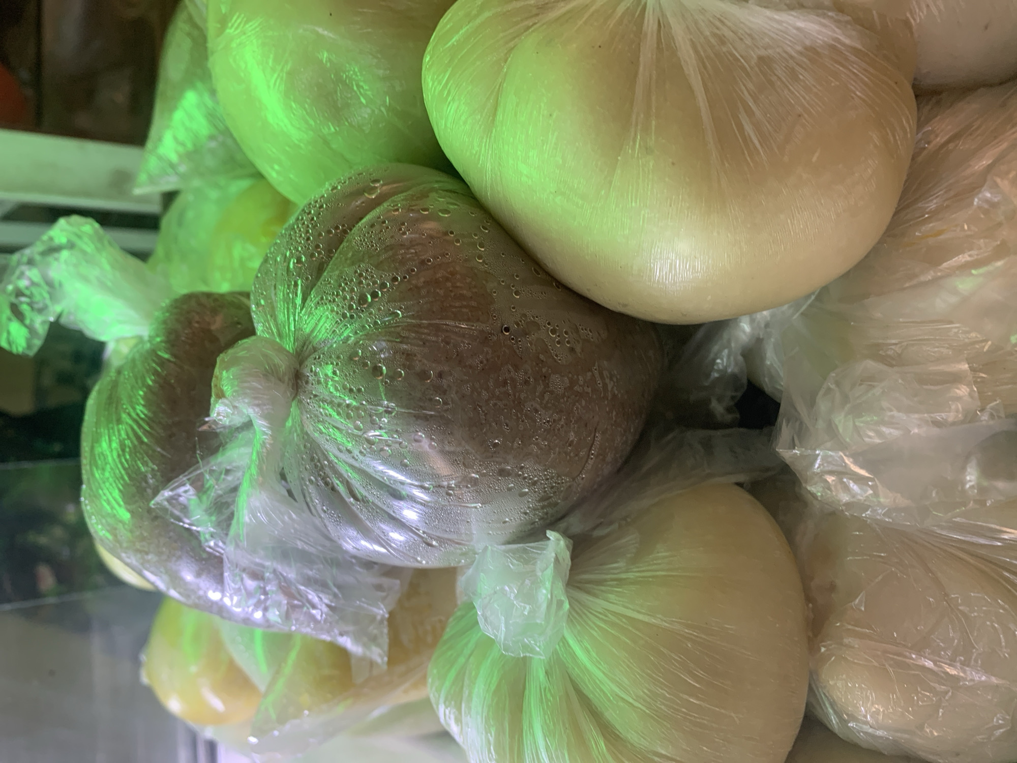 A close-up, high-angle shot captures a dense cluster of various food items, presumably Nigerian staples such as pounded yam or fufu, encased within clear plastic bags. The items are irregularly shaped, primarily spherical or ovoid, and stacked in an ad hoc manner. Most visible items are a pale, off-white to yellowish color, while one prominent item centrally located and slightly elevated exhibits a darker, reddish-brown hue. All plastic bags show signs of significant internal condensation, visible as small water droplets clinging to the inner surface, particularly pronounced on the darker item. A strong, artificial green light source illuminates the scene, casting a distinct verdant glow and reflections across the translucent plastic surfaces. No individuals are present. The background is dark and out of focus, suggesting an indoor commercial setting, likely a food stall or market display in the New Stadium area of Nigeria.