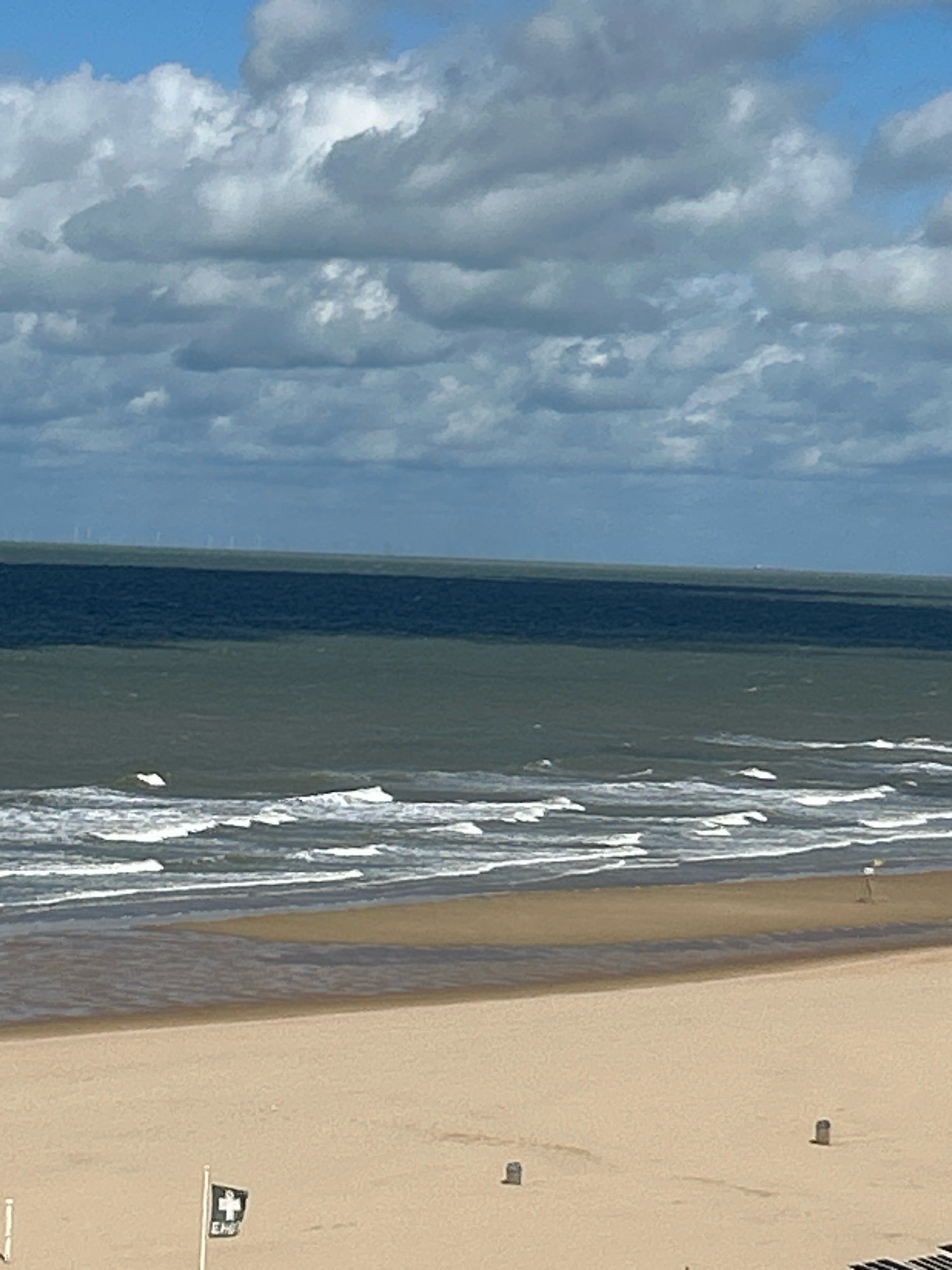 This is an outdoor, eye-level, medium shot of the beach and sea in Ostend, Belgium, under a partially cloudy sky. The image captures a wide expanse of sandy beach in the foreground, transitioning to the choppy sea with white-capped waves. In the distance, a dark line of trees or shrubbery forms a horizon, beyond which, faint wind turbines are visible. There are a few small, dark cylindrical objects scattered on the beach, possibly posts or small structures. To the left, a flag with a white cross on a black background and some text is planted on a pole. The overall scene is tranquil, with no people immediately visible in the frame.