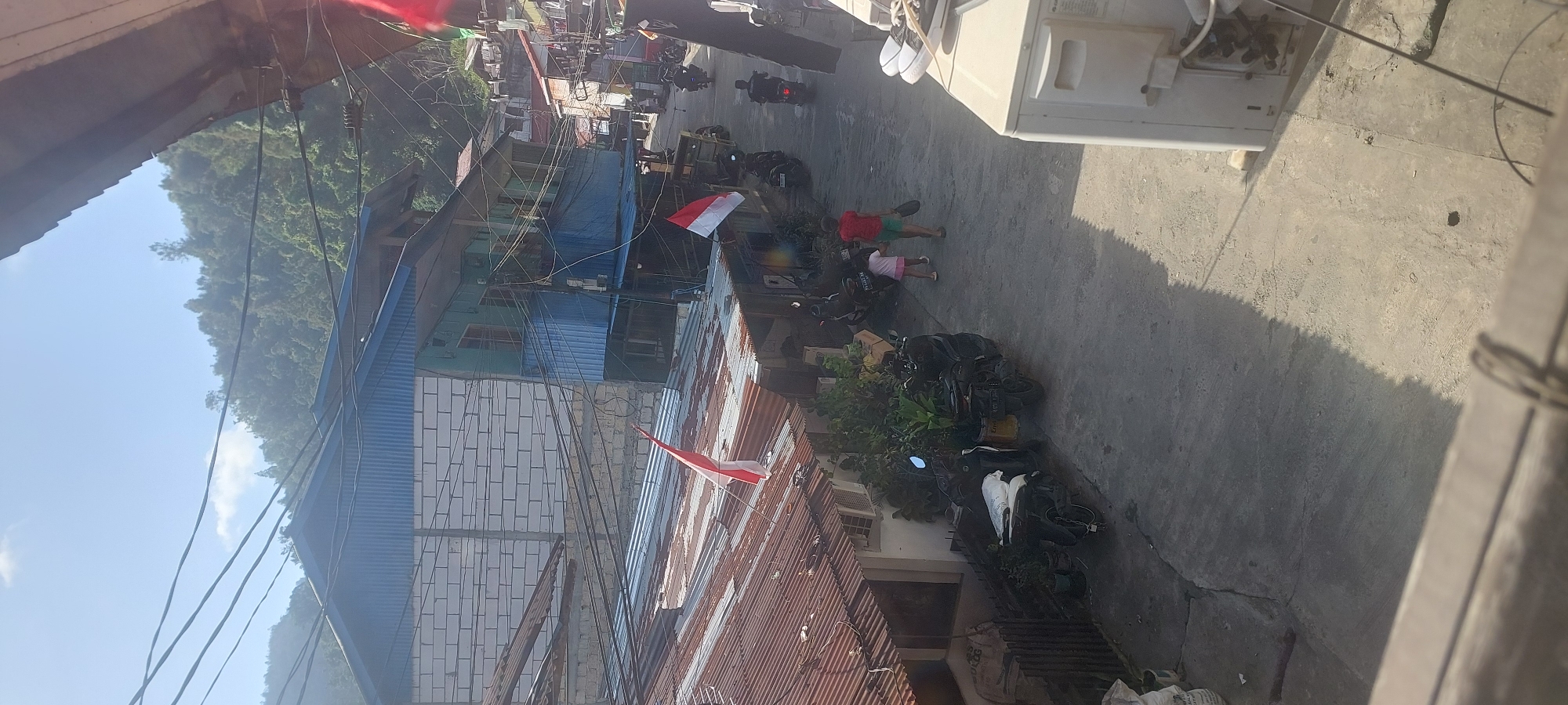 The image depicts a street scene in a densely populated area, likely a residential neighborhood in Jayapura, Indonesia. In the foreground, a concrete surface with cracks and some debris is visible. To the right, the side of a white air conditioning unit is prominently featured, with its external components and wiring exposed. A wooden railing runs across the bottom of the frame, obscuring some of the lower details.
Further into the scene, a narrow street is lined with buildings and parked motorcycles. Several motorcycles, some appearing to be covered with bags or tarps, are parked along the left side of the street. A building with a corrugated metal roof, showing signs of rust, occupies the middle ground. A red and white flag, potentially the Indonesian flag, is affixed to a pole on this roof. Adjacent to this building is another structure with a blue corrugated metal roof and white brick walls. A two-story building with a light blue facade and dark blue trim is situated behind the first building.
Two children are walking away from the viewer down the street. One child, wearing a red shirt and green shorts, is holding a black object, possibly a shoe. The other child, in a pink shirt and shorts, is walking beside them. Other motorcycles and buildings are visible further down the street, with some individuals on motorcycles riding away from the camera. The background shows a lush, tree-covered hillside under a bright, partly cloudy sky. The overall impression is of a bustling urban environment with a mix of residential and commercial structures, typical of a developing city.
