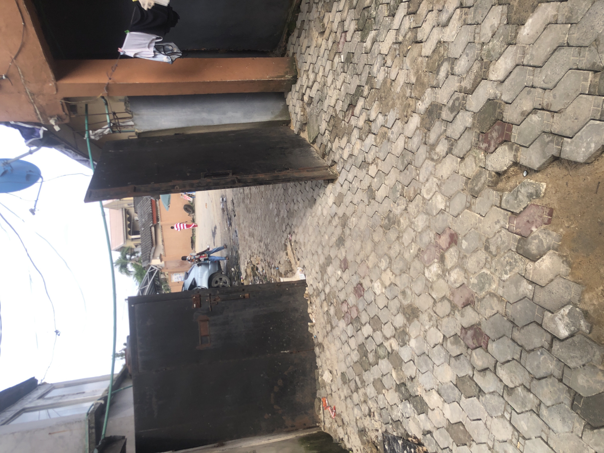 The image depicts an outdoor scene in Ado, Nigeria, characterized by a courtyard paved with interlocking hexagonal bricks. On the left, a large, dark metal gate is partially open, revealing a silver car parked behind it. To the right of the car, a person wearing blue jeans and a dark top is standing, holding a baby. Further into the courtyard, a person in a red and white striped outfit is walking away from the camera.
Dominating the right side of the image is a section of an orange building with an open doorway where laundry, including a white bra and black undergarments, is hanging on a clothesline. A blue satellite dish is visible on the roof of a building in the background, along with power lines. The environment appears somewhat unkempt, with debris scattered on the ground and the paving stones showing signs of wear and dirt. The sky is overcast.