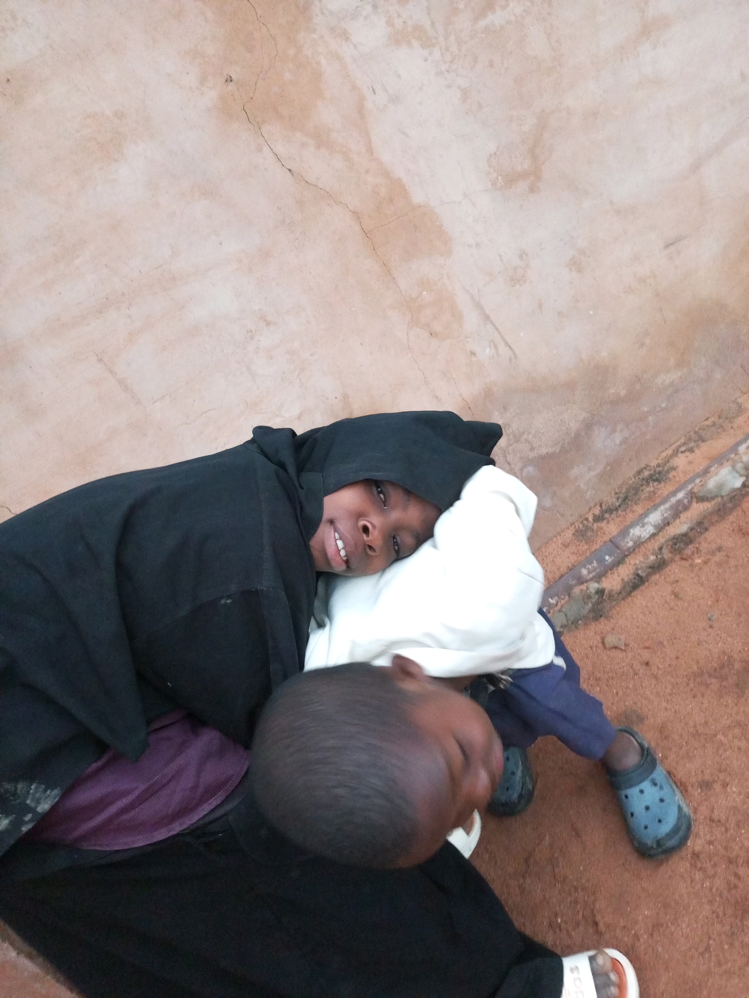 The image shows two children lying down, possibly on the ground, with one child's head resting on a white pillow or folded fabric. The child closer to the camera is wearing a black hooded garment, and a purple garment is visible underneath. The other child is wearing blue pants and blue clogs. The background appears to be a textured, light-colored wall with some cracks. The ground beneath the children is a reddish-brown dirt. This scene is likely located in Tempure, Nigeria, but the specific context or activity is not discernible from the image alone.