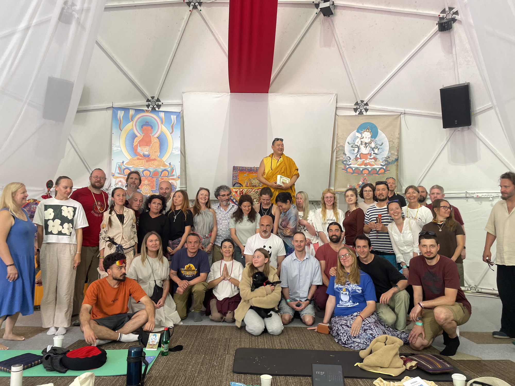 The image shows a group of approximately 40 people posing for a group photograph indoors, likely in Mekića Kula, Montenegro. A person in saffron robes, possibly a monk or spiritual leader, stands on a raised platform in the background, holding a small white rectangular object. Behind him are two Tibetan-style religious paintings flanking a red and white drape. The people in the foreground are a mix of ages and ethnicities, and are either standing, kneeling, or sitting on the floor. In the foreground, there are yoga mats, water bottles, books, and other personal items scattered around. The location appears to be a temporary or semi-permanent structure with a domed ceiling and white fabric walls. A black speaker is mounted on the wall in the upper right.