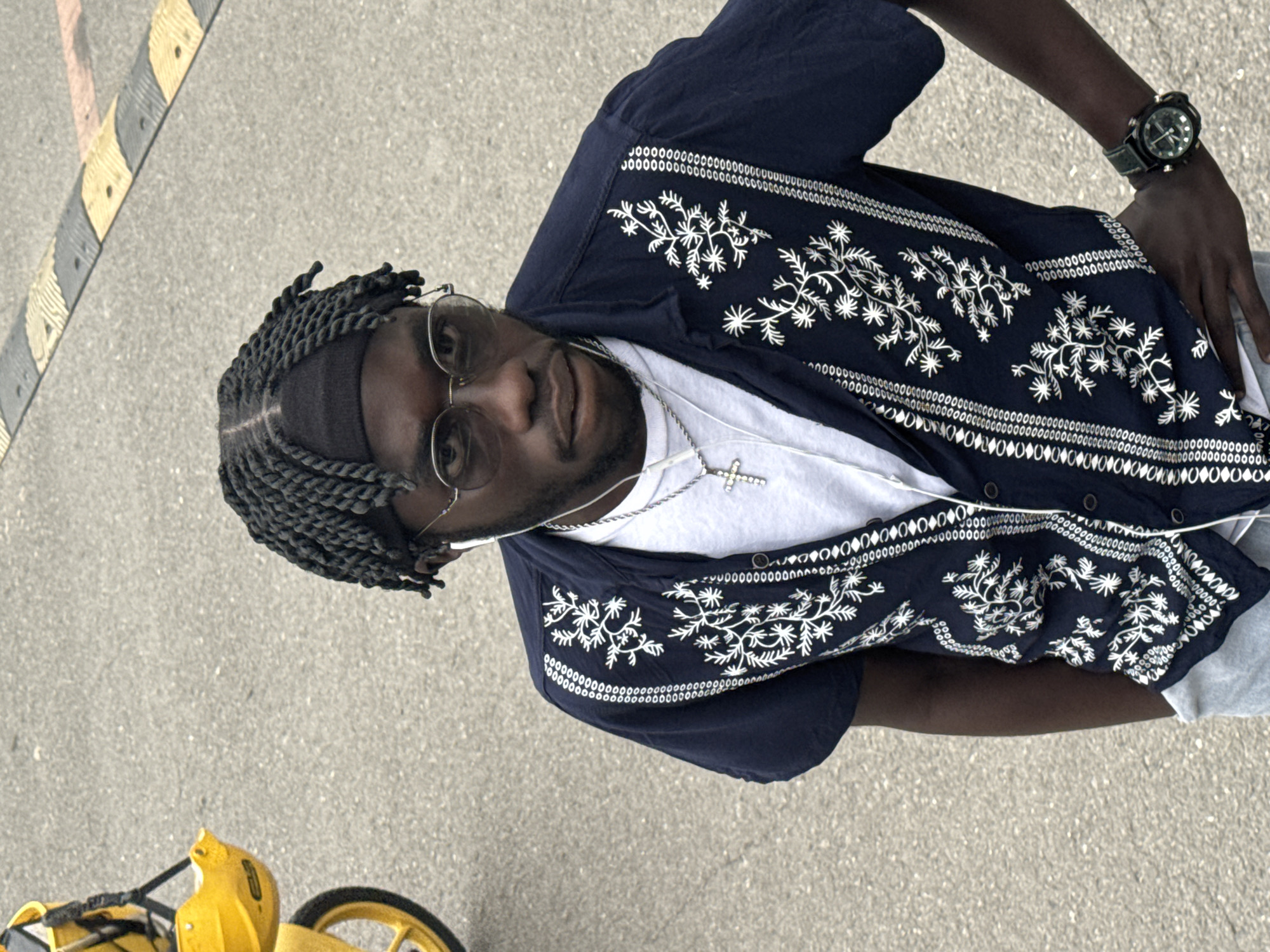 A medium-shot of a dark-skinned man is taken from an elevated perspective. The man has dark hair styled in braids and is wearing silver, round-rimmed glasses. He is also wearing a white t-shirt under a dark blue button-down shirt with ornate white embroidery. A silver chain necklace with a cross pendant is visible on his chest. On his left wrist, he wears a large green watch.
The man is standing on an asphalt surface, and a yellow scooter is partially visible in the lower-left corner of the frame. The asphalt surface has a gray and yellow striped marking running along the left edge of the frame.
The location is Zhongtun, China.