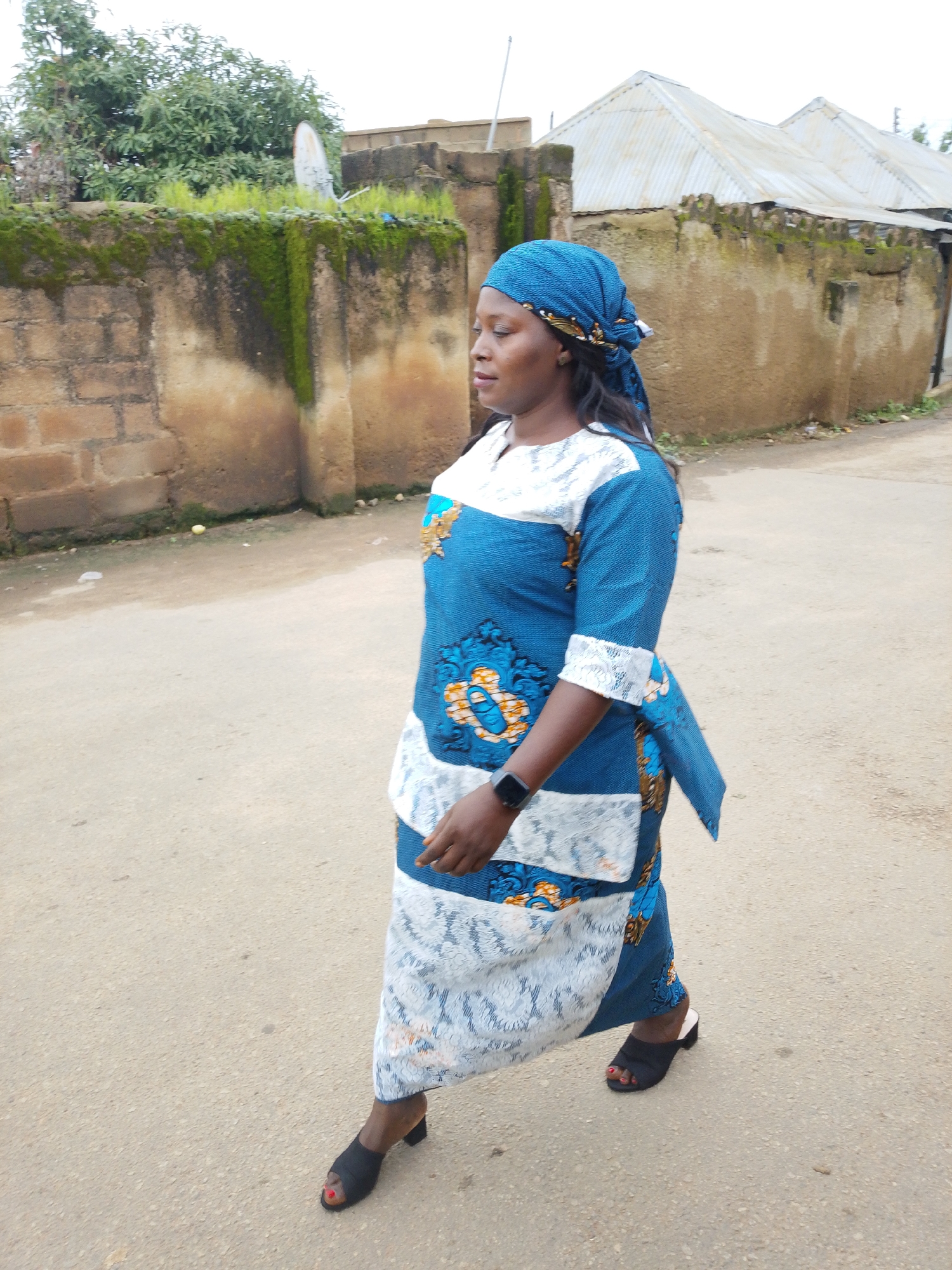 In Jos, Nigeria, a woman walks on a dirt road. She is wearing a blue and white patterned dress, a blue headscarf, and black open-toed heels. Her dress has a blue base with white lace trim and floral patterns in blue, orange, and yellow. She has a smartwatch on her left wrist. The background features a textured, light brown brick wall covered in moss, with green foliage growing on top. A white satellite dish is visible on the wall. Beyond the wall, there are corrugated metal roofs of buildings and some trees. The road surface is dusty and uneven, with scattered debris. The overall scene depicts an outdoor urban environment in Jos, Nigeria, with a focus on the woman in motion.