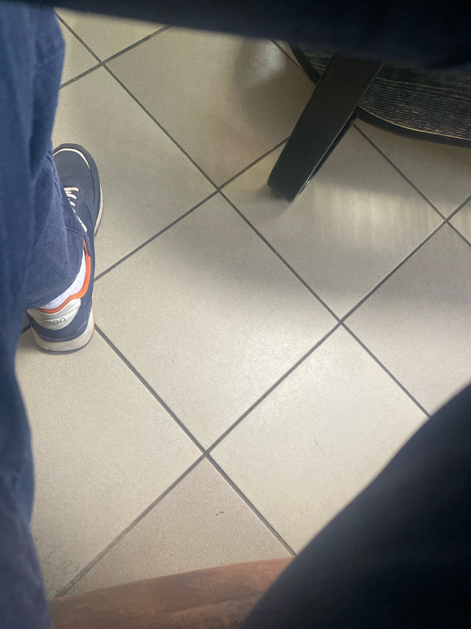 The image shows a close-up view of a person's legs and feet on a tiled floor, likely indoors. The person is wearing blue pants and a blue and white sneaker with orange accents. The floor is made of light-colored square tiles with dark grout lines, laid in a diamond pattern. A black chair leg is visible in the upper right portion of the image. The lighting suggests a daytime setting, with shadows cast on the floor. The specific location in Minsk, Belarus cannot be determined from the visual information provided.
