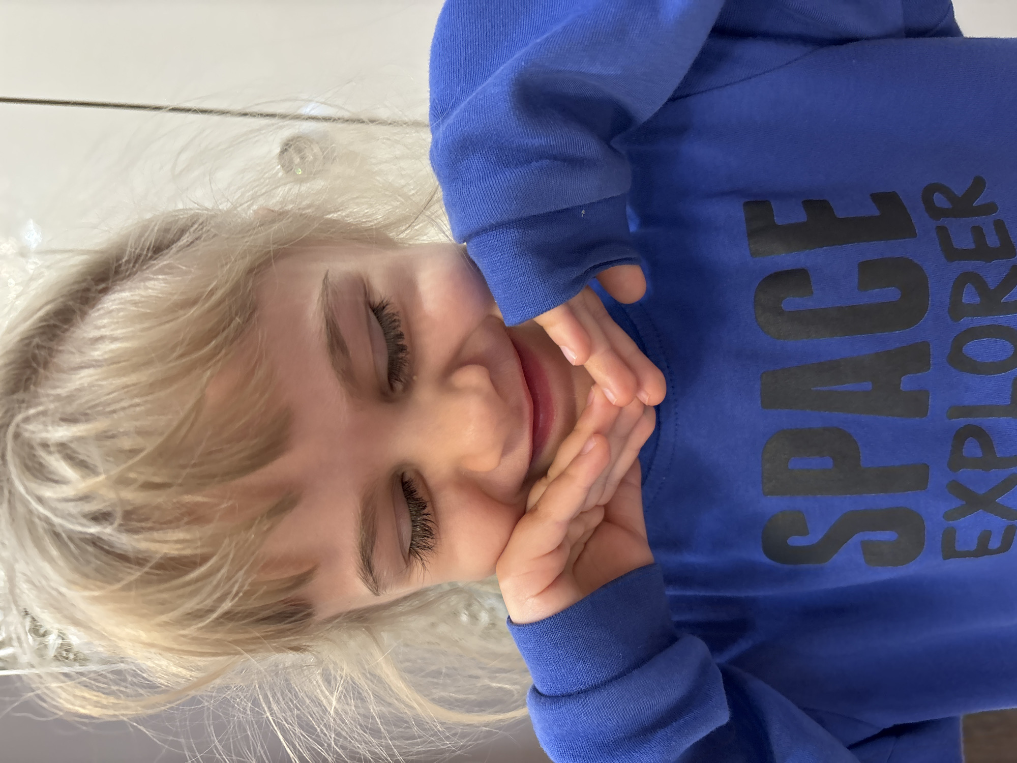 A close-up image shows a young child with blonde hair wearing a blue long-sleeved shirt with the words "SPACE EXPLORER" printed in black. The child's face is tilted, with their eyes closed and hands gently touching their cheeks. The background is blurred and shows a white surface.