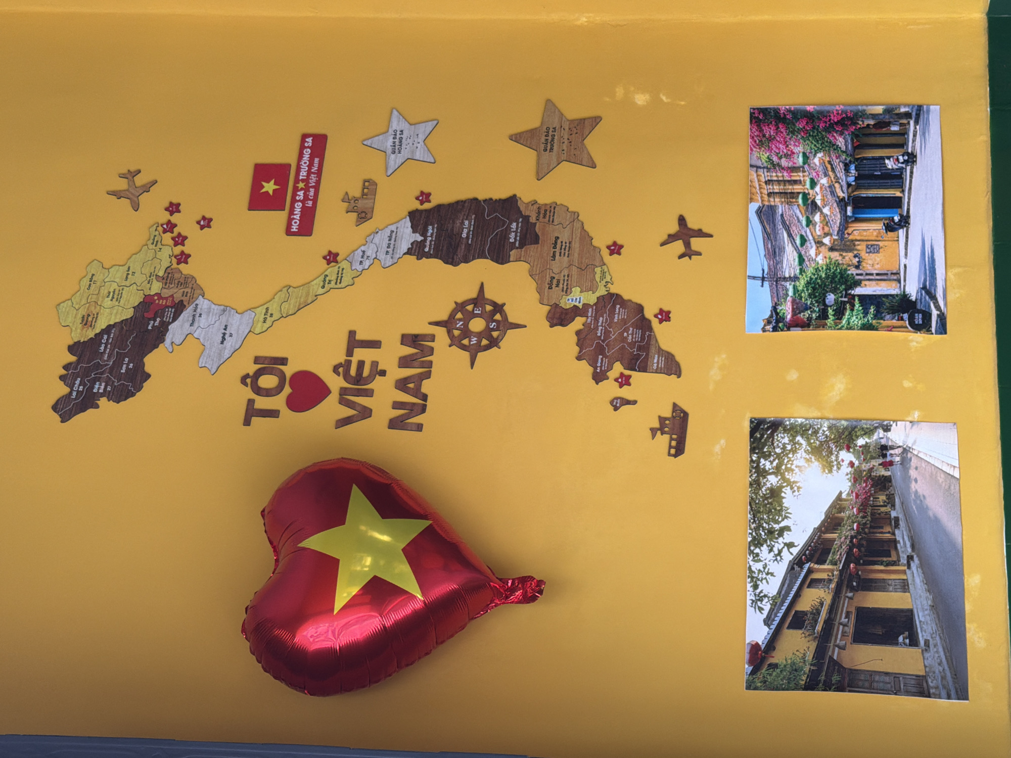 The image depicts a yellow wall decorated with a map of Vietnam made of wood cutouts, a red heart-shaped balloon with a yellow star, and two photographs of street scenes. The map of Vietnam features various cities and regions labeled with text. A wooden cutout of an airplane is positioned above the map, and a boat cutout is located to the right of the map. Several red stars and two larger wooden stars, one labeled "QUẦN ĐẢO HOÀNG SA" and the other "QUẦN ĐẢO TRƯỜNG SA," are also attached to the wall. The photographs show picturesque street views of what appears to be Hoi An, Vietnam, characterized by yellow buildings, flowering trees, and lanterns. The left photograph shows a street with yellow buildings and a few people walking, while the right photograph displays a similar street with more vibrant pink flowers and parked motorbikes. The overall setting is an indoor environment with a bright yellow wall. The description of the location in the city of Nha Trang, Vietnam, is not supported by any visual evidence in the image.