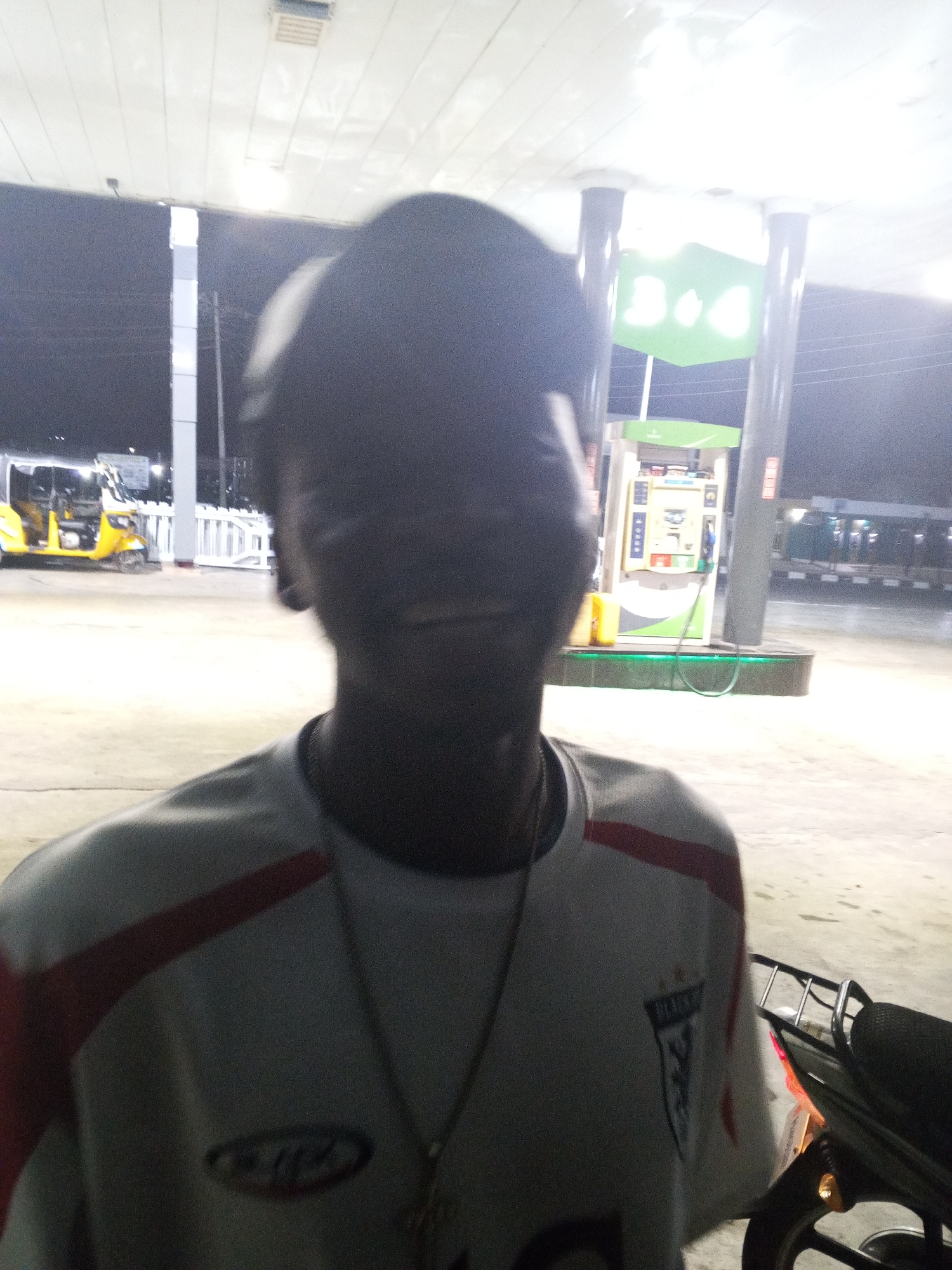 At a gas station in Lokoja, Nigeria at night, a person with an obscured face is standing in front of the camera, wearing a white t-shirt with red stripes on the shoulders and a necklace with a cross pendant. In the background, there's a yellow tricycle, gas pumps, and a motorcycle is partially visible on the right side of the frame. The lighting is bright, possibly from the gas station's overhead lights.