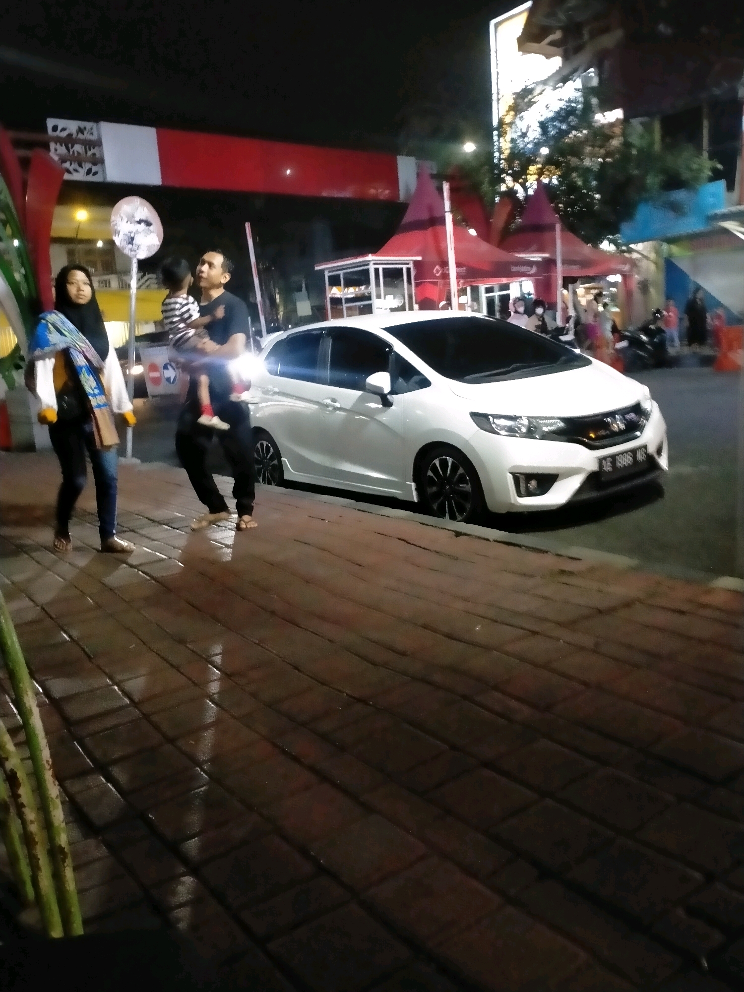 The image depicts a street scene at night in Batu, Indonesia, likely near a food stall or market area as indicated by the red awnings and illuminated tents in the background. A white hatchback car, possibly a Honda Jazz based on its styling and license plate which reads "AE 1886 WS", is parked along the sidewalk. The sidewalk itself is paved with rectangular bricks and appears wet, reflecting the ambient light.
In the foreground, to the left of the car, a man is holding a young child. The man is wearing a dark t-shirt and dark pants, while the child is in a striped shirt. To the left of them, a woman wearing a hijab and a light-colored jacket with a blue and yellow scarf is walking. Her dark pants and sandals suggest she is also on the sidewalk.
The background shows a vibrant street environment with various stalls and buildings illuminated by artificial lights. Trees are visible, suggesting a possibly green area within the city. The overall atmosphere is lively and suggests a commercial or recreational area. The presence of the car, people, and stalls indicates a public space in Batu.