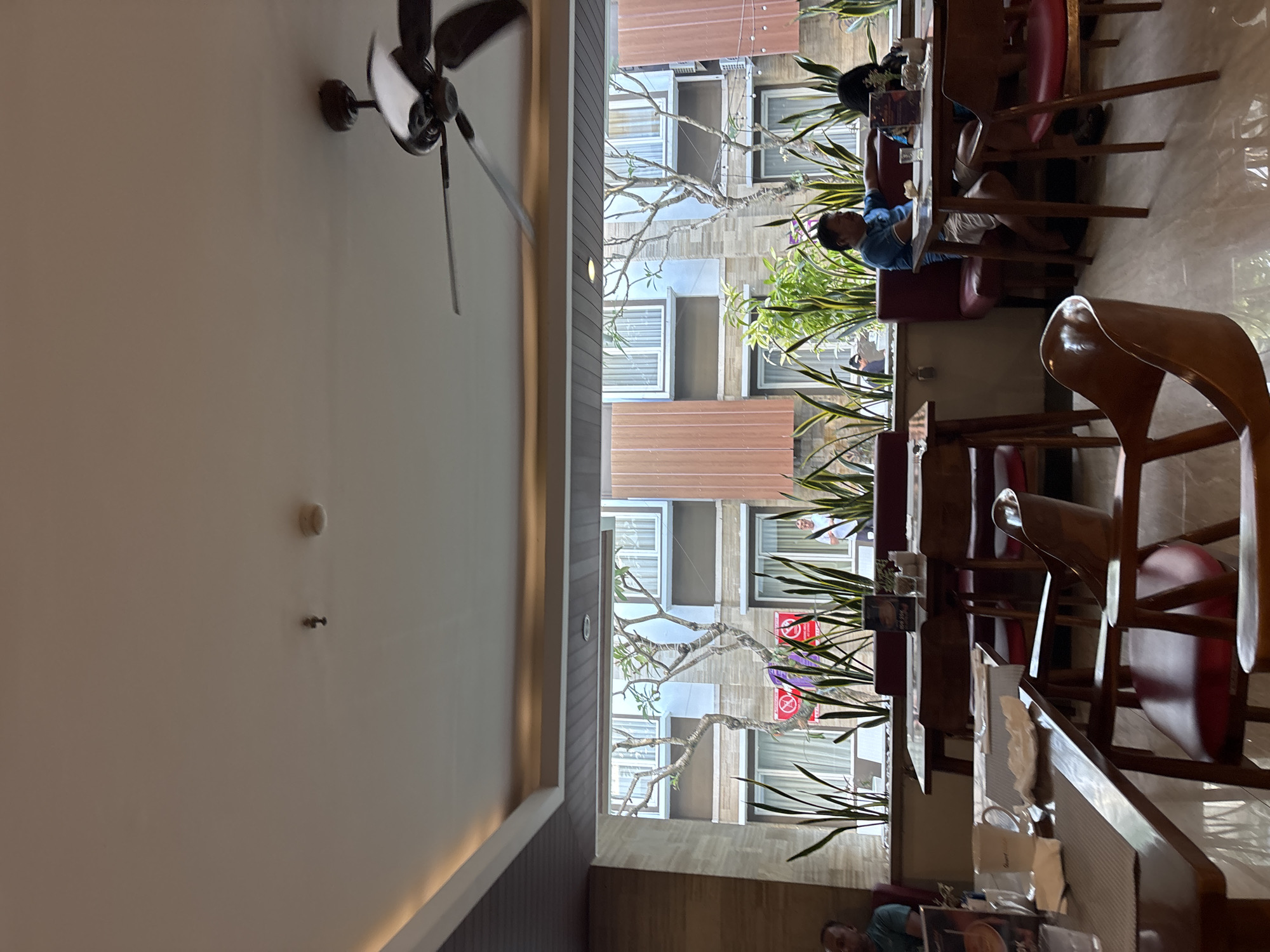 The image captures an indoor view of a restaurant or cafe with an open-air ambiance, looking out towards a courtyard or building facade. The interior features wooden tables and chairs with red upholstery, set on a polished floor. A ceiling fan is visible, along with lighting fixtures and a smoke detector.
Through a large open window or partition, a courtyard is visible with numerous tall, green plants. Beyond the courtyard, there is a multi-story building with a repeating pattern of windows and balconies. The building's facade appears to be a mix of light blue and brown tones, with some wooden paneling. Trees with bare branches are also visible in the courtyard, suggesting a tropical or subtropical climate.
Several patrons are seated at tables. On the left side, a person is partially visible sitting at a table. In the middle, a man in a blue shirt and white shorts is seated at a table, leaning on his arm. To his right, another person is seated, partially obscured. In the background, a person in a white shirt is standing near the building.
The overall impression is of a dining establishment that embraces natural light and a connection with the outdoors. The setting appears to be a modern hotel or resort in Denpasar, Indonesia, given the architectural style, climate, and typical amenities.