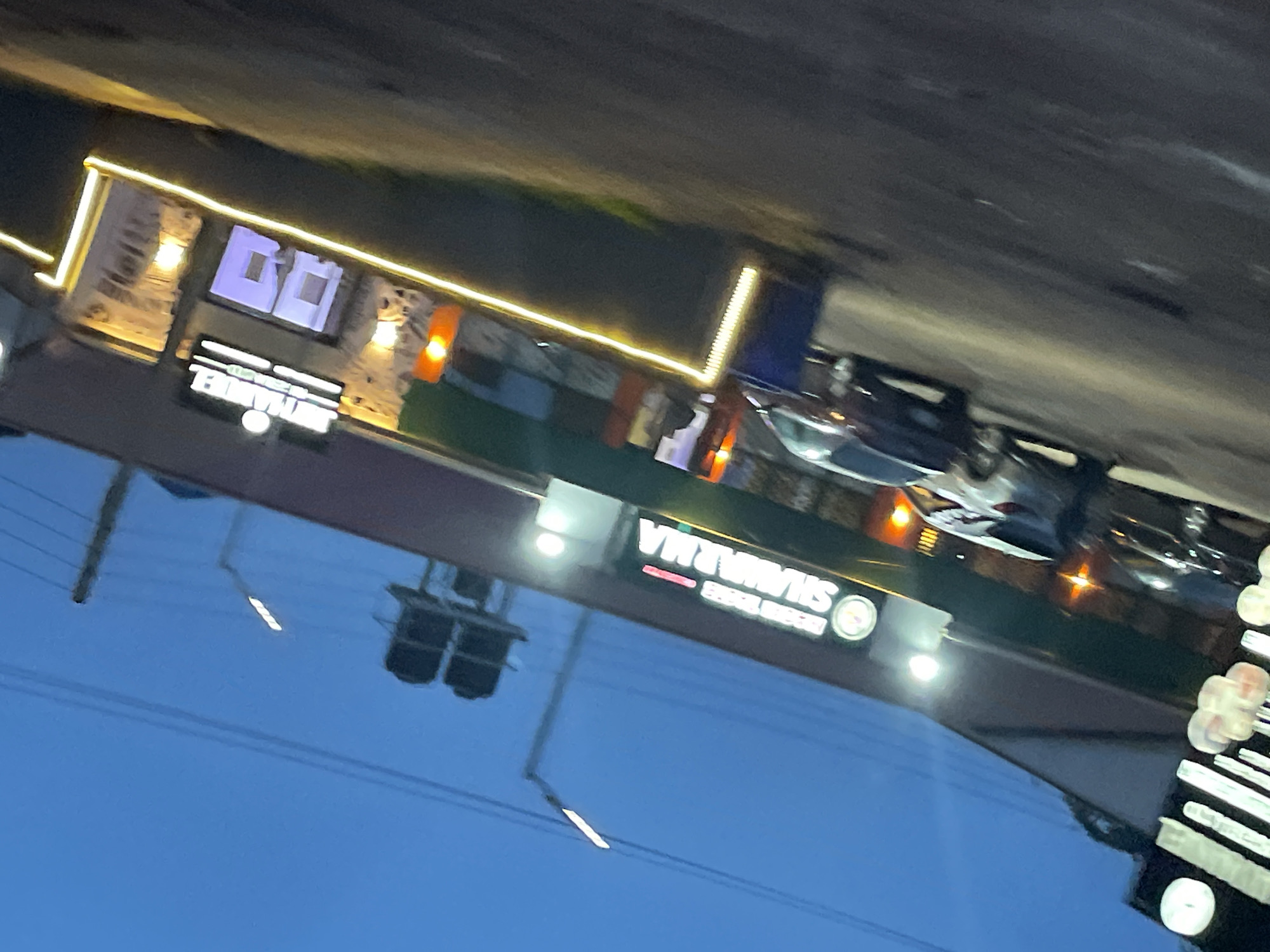 The image shows a street scene in Jos, Nigeria, at dusk or early evening. A building with a prominent illuminated sign reading "Shawarma" is visible, suggesting a food establishment. Several cars are parked or driving in front of the building, and the street is lined with lights. The sky is a deep blue, indicating a transition from day to night. The overall impression is that of a commercial area in an urban setting.