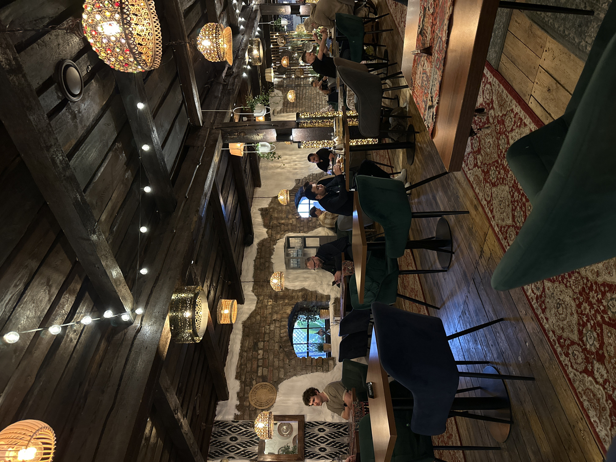 The image depicts the interior of a restaurant. The walls are textured with brick and plaster. Patrons are seated at tables, some interacting with electronic devices, others in conversation. The seating consists of velvet chairs in colors of emerald, navy, and ash. Several light fixtures adorn the ceiling, a mix of decorative lanterns and string lights, along with rustic wooden beams. The floor is wooden. A rug with a decorative pattern runs along the side of the table area. Small windows are visible in the brick walls. The location is in the city of Unknown, Unknown.