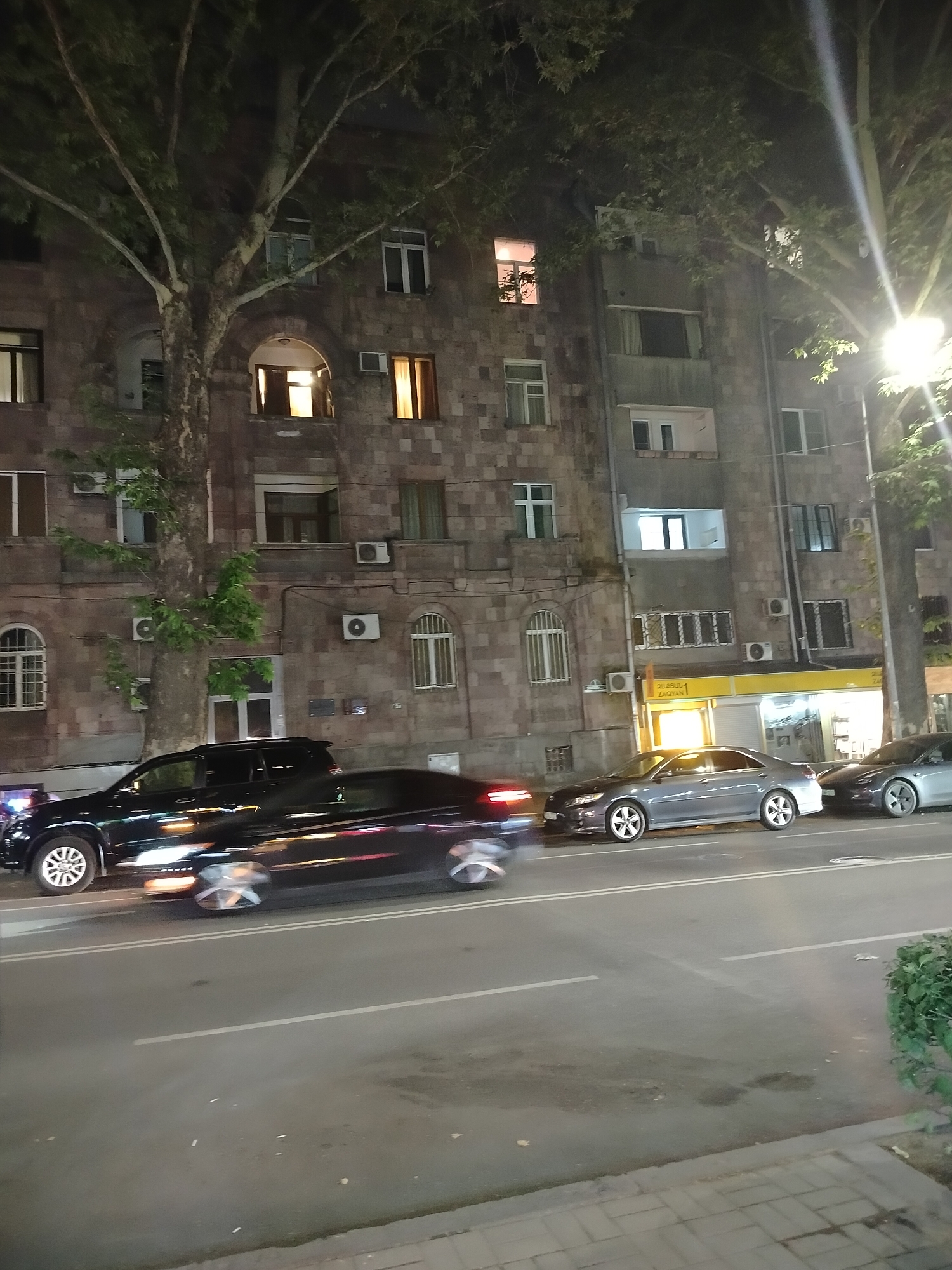 The image shows a street scene at night in Yerevan, Armenia. A multi-story building is on the right side of the image with trees obscuring some parts. Several cars are parked along the curb, and one car is moving, creating a light blur. Above the cars, on the first floor of the building, a storefront is visible with yellow signage. The building has several windows, some lit, and numerous air conditioning units affixed to the facade. The lighting is provided by street lamps and building lights. A sidewalk and paved area are in the foreground.