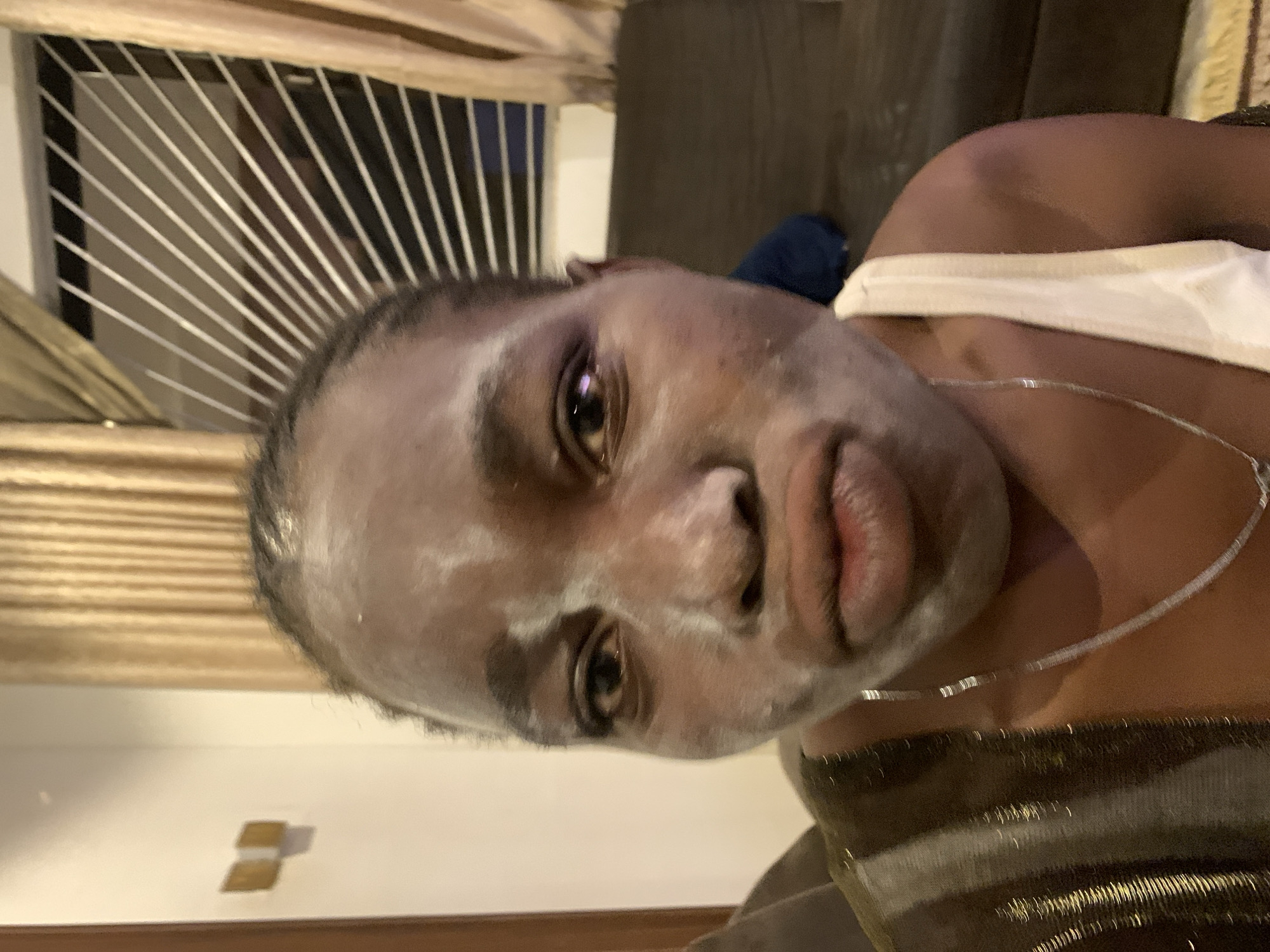 A medium shot depicts a woman with a light-colored facial mask. She is wearing a silver chain necklace and a white tank top. The background features vertical bars behind her head and a brown couch. Jos, Nigeria is not discernible from the image.
