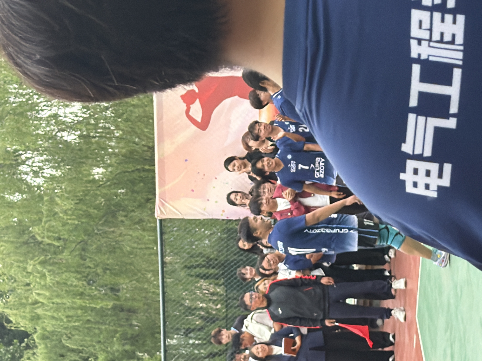 The image shows a group of people standing on what appears to be an outdoor sports court. There is a banner visible in the background with a red silhouette of a volleyball player. The people in the group are of mixed ages and are wearing a variety of clothing. Some individuals are wearing blue athletic jerseys with white lettering. A person in the foreground is wearing a blue shirt with Chinese writing, possibly indicating the word 'engineering'. The environment includes a green court surface and trees visible in the background, suggesting a park or outdoor venue. No specific information to pinpoint Guta is readily available in the image.