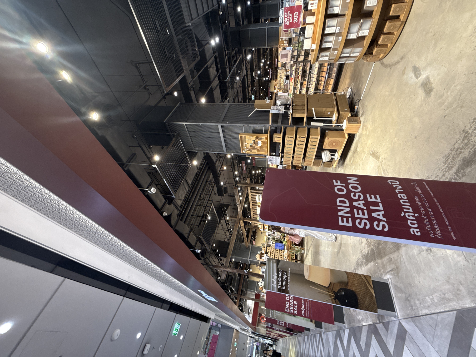 The image shows the interior of a large retail store, possibly a department store or a lifestyle store, in Bangkok, Thailand. The store has a modern industrial aesthetic with exposed ceilings, metal shelving units, and wooden display shelves. There are several signs visible, including one that reads "END OF SEASON SALE" with "30%" discount. These signs appear to be in both English and Thai. The store is well-lit with a combination of overhead fluorescent lights and spotlights. In the foreground, there are tall red banners with white text, advertising the sale. Further into the store, shelves are stocked with various products, including boxes, decorative items, and what appear to be kitchenware or home goods. A few people are visible, including a woman in the background walking away from the camera. The floor is concrete. The overall impression is of a busy retail environment during a sale event.