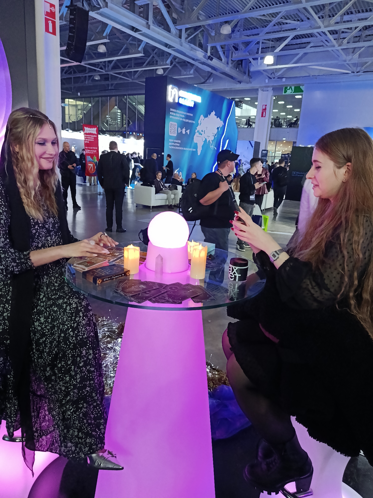 The image captures a scene from an event, possibly a convention or trade show, taking place indoors in Khimki, Russia. Two women are the central figures, seated at a round glass table supported by a neon pink pillar.
The woman on the left, fair-skinned with long wavy blonde hair adorned with a beaded headpiece, wears a dark floral dress with a sheer black scarf. She is actively engaged with a spread of tarot cards on the table, her hands lightly touching them. On the right, another woman with fair skin and long brown hair is seated, dressed in black. She holds an object in her hands, possibly a phone, seemingly showing it to the tarot reader.
On the table, besides the tarot cards, are several lit battery-operated candles, a clear crystal, and a white spherical light fixture, adding a mystical element to the setup. There's also a dark container with white text and a coffee mug.
The background reveals a busy exhibition hall with a mix of people in business attire. Exhibition booths and large displays are visible, including one with a world map and Russian text. There are also lounge areas with white sofas. Overhead, the exposed structural framework of the building is visible, along with speakers suspended from the ceiling. The lighting is a mix of natural light and artificial lighting from the convention center, with the pink glow from the table contrasting against the overall ambiance.