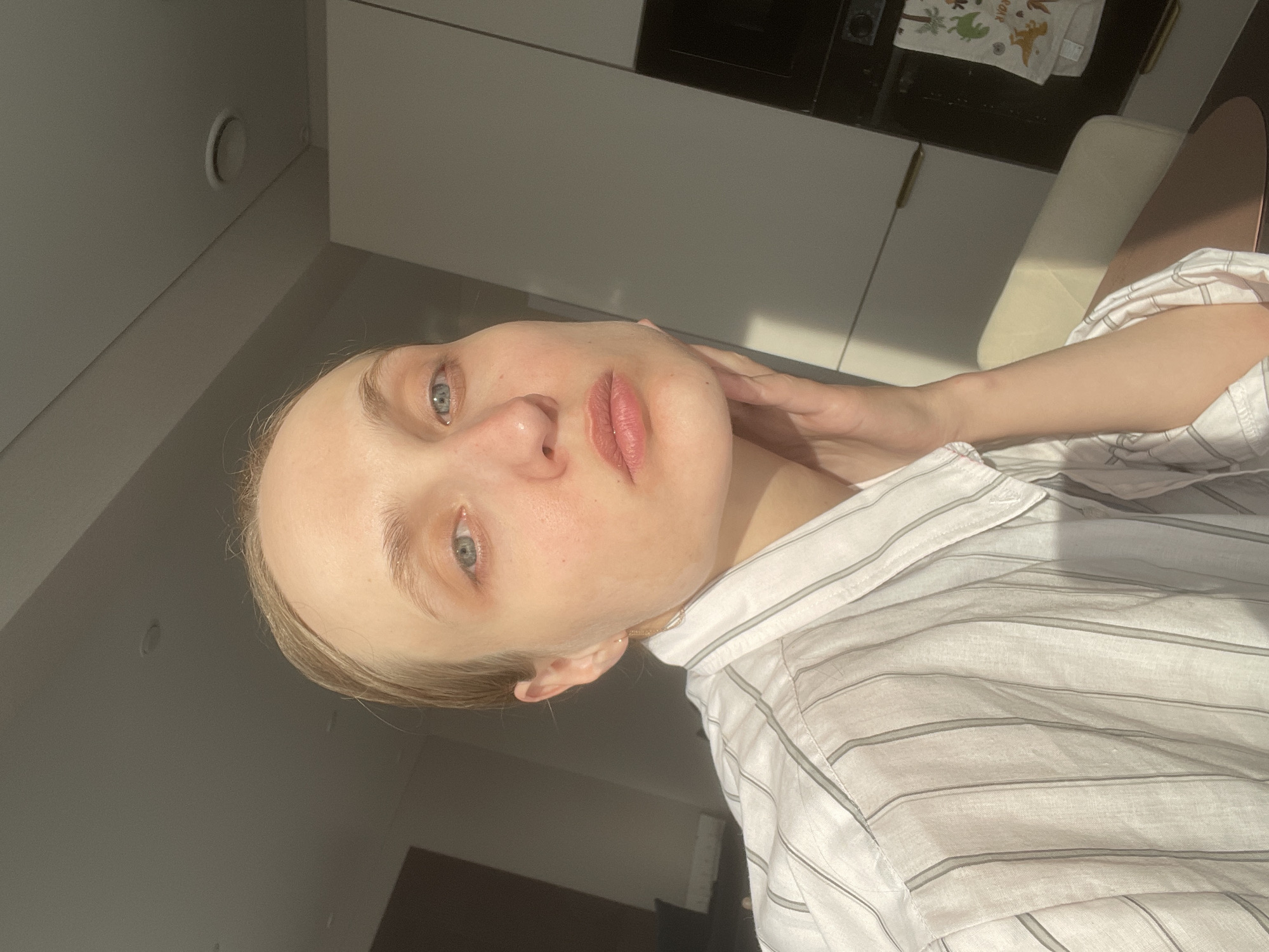 The image presents a selfie of a light-skinned individual with blonde hair and blue eyes. The person is wearing a white striped shirt and is positioned at an angle, with their right hand touching their neck. The setting appears to be indoors, likely a kitchen, given the presence of cabinets and a kitchen towel visible in the background. The lighting suggests daytime. There is no information to confirm that this image was taken in Sepych, Russia.