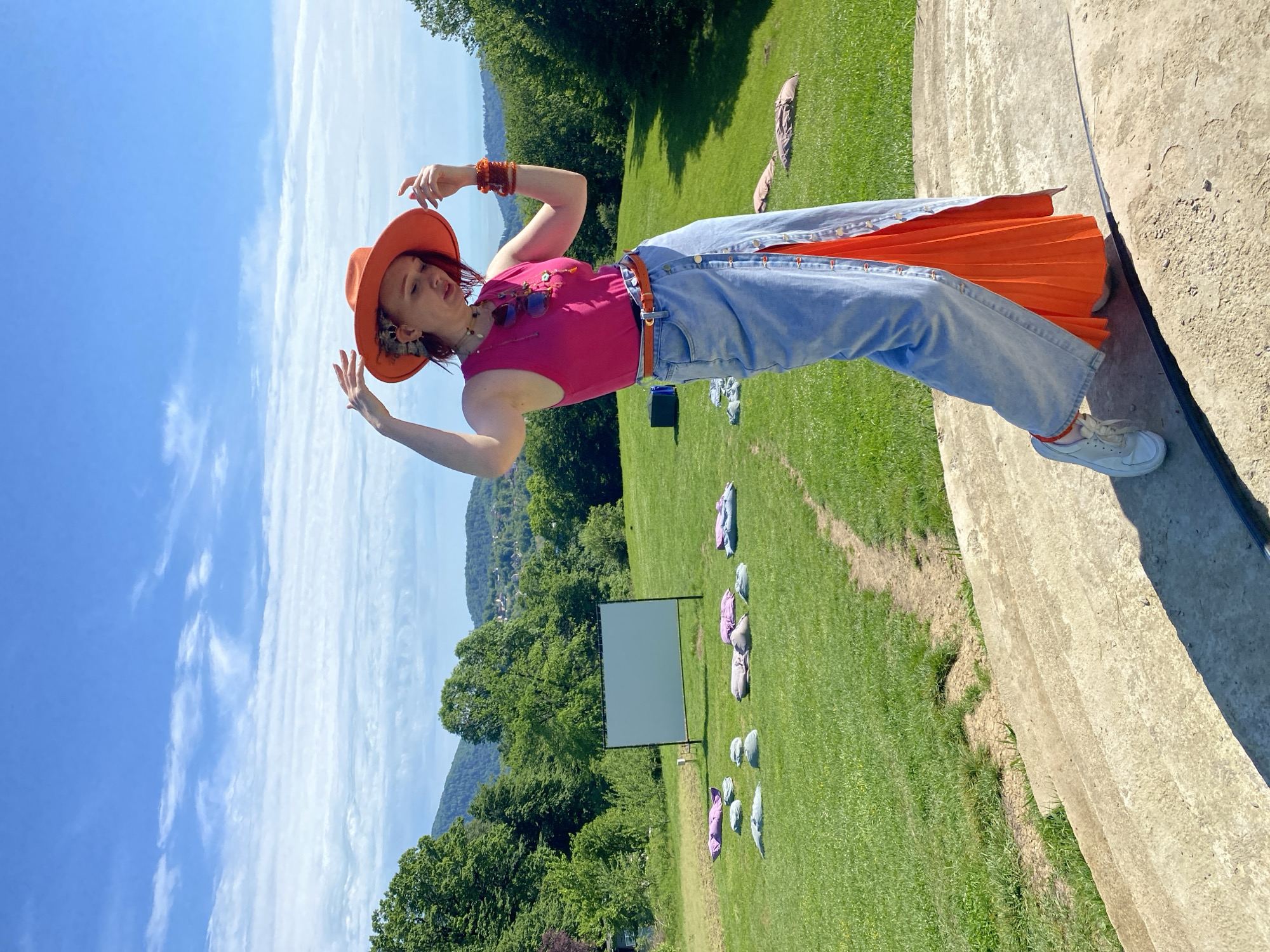 The image depicts a woman standing on a concrete structure overlooking a grassy field. She is wearing an orange hat, a pink tank top, light blue jeans with an orange pleated skirt underneath, and white sneakers. She has her arms raised with her hands open.
In the background, there is a large outdoor screen set up in the field, and blankets are scattered around the grass in front of it. Beyond the field, there are trees and a body of water or sky. The sky is blue with wispy clouds.
There is no clear indication of this scene being in the city of Прогресс, Russia.