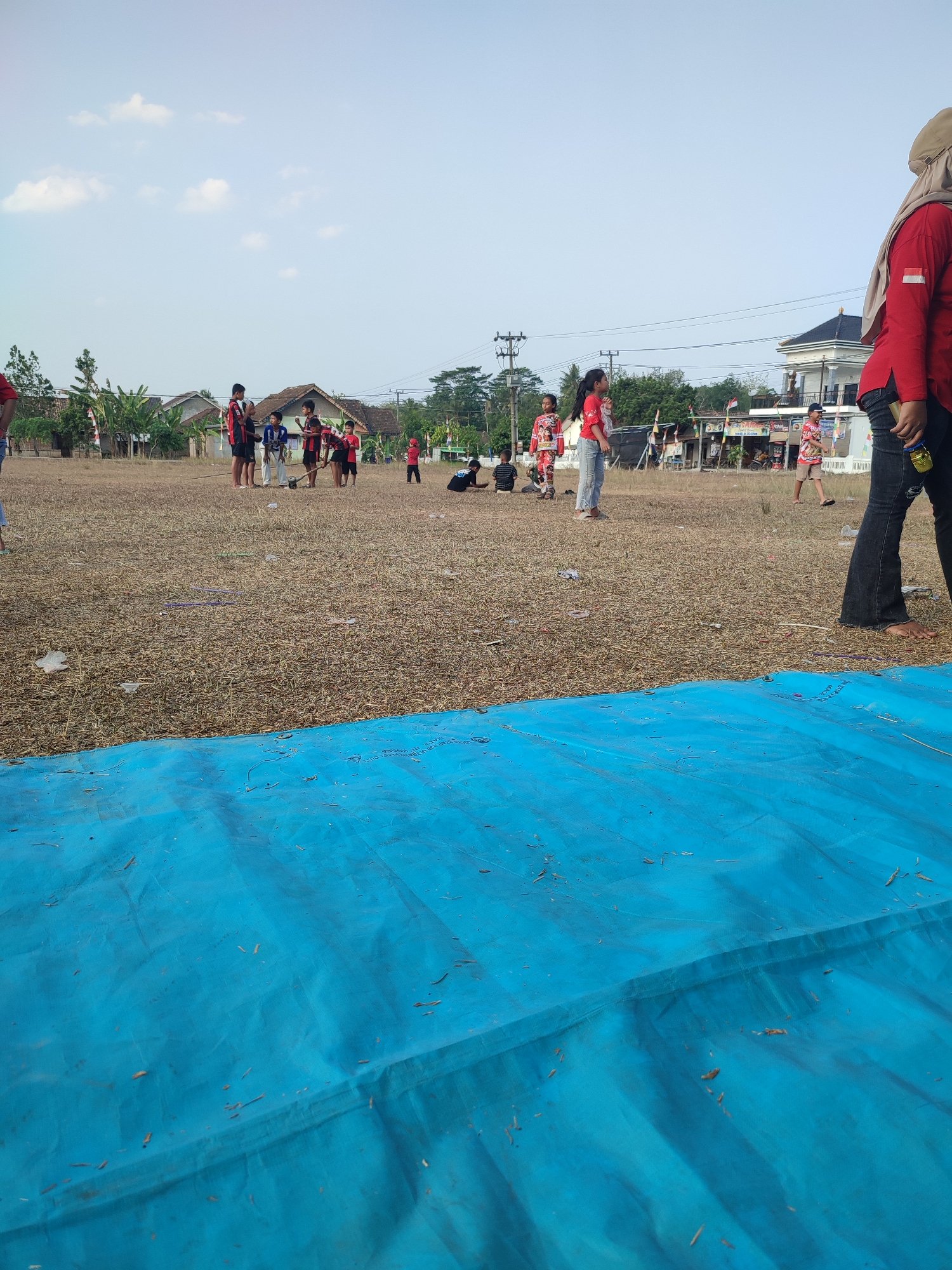 In the city of Nusa Jaya, Indonesia, a group of children are gathered in a dry, grassy field under a clear sky. The field is sparsely populated with dry grass and some scattered debris. In the foreground, a large, bright blue tarp covers a significant portion of the ground. In the background, several houses with red-tiled roofs are visible, along with some trees and utility poles.
A woman wearing a red long-sleeved shirt and a hijab is standing on the right side of the image, facing away from the camera. She is holding a yellow object in her right hand. A group of children, mostly boys, are in the middle ground, some wearing red shirts with white accents. They appear to be engaged in an activity, possibly a game or a demonstration, as some are holding sticks. One boy is wearing a blue outfit. To the left of this group, a few children are also standing, and further to the left, a person in a red shirt is partially visible. On the far right, a young child in a red outfit is standing. In the background, beyond the field and houses, is a glimpse of a larger white building. The overall scene suggests an outdoor event or gathering in a rural or semi-rural setting.