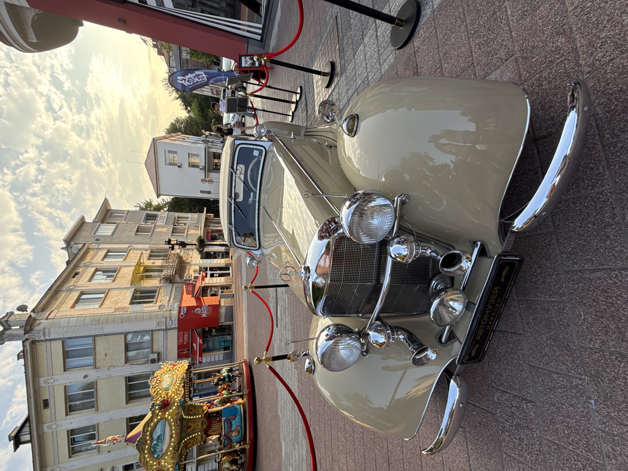 The image shows a vintage beige convertible Mercedes-Benz car parked on a cobblestone street in Plovdiv, Bulgaria. The car is the main subject, positioned in the foreground and angled towards the viewer. Its chrome details, large headlights, and grille are prominent. Behind the car, a carousel with colorful horses and a canopy is visible on the left, suggesting a public square or pedestrian zone. Buildings with light-colored facades, some with balconies and arched windows, line the street on the left and in the background. On the right, a modern red and white structure with a tall, curved element and a blue banner is present. Several people are walking in the background, and red velvet ropes on stanchions demarcate the area around the car. The sky is partly cloudy with a warm sunset glow, casting a soft light on the scene. The license plate of the car reads "АВТОМУЗЕЙ" and "МАРКОВО," indicating it is from the Auto Museum in Markovo, a village near Plovdiv. The overall impression is of a classic car display in an urban environment.