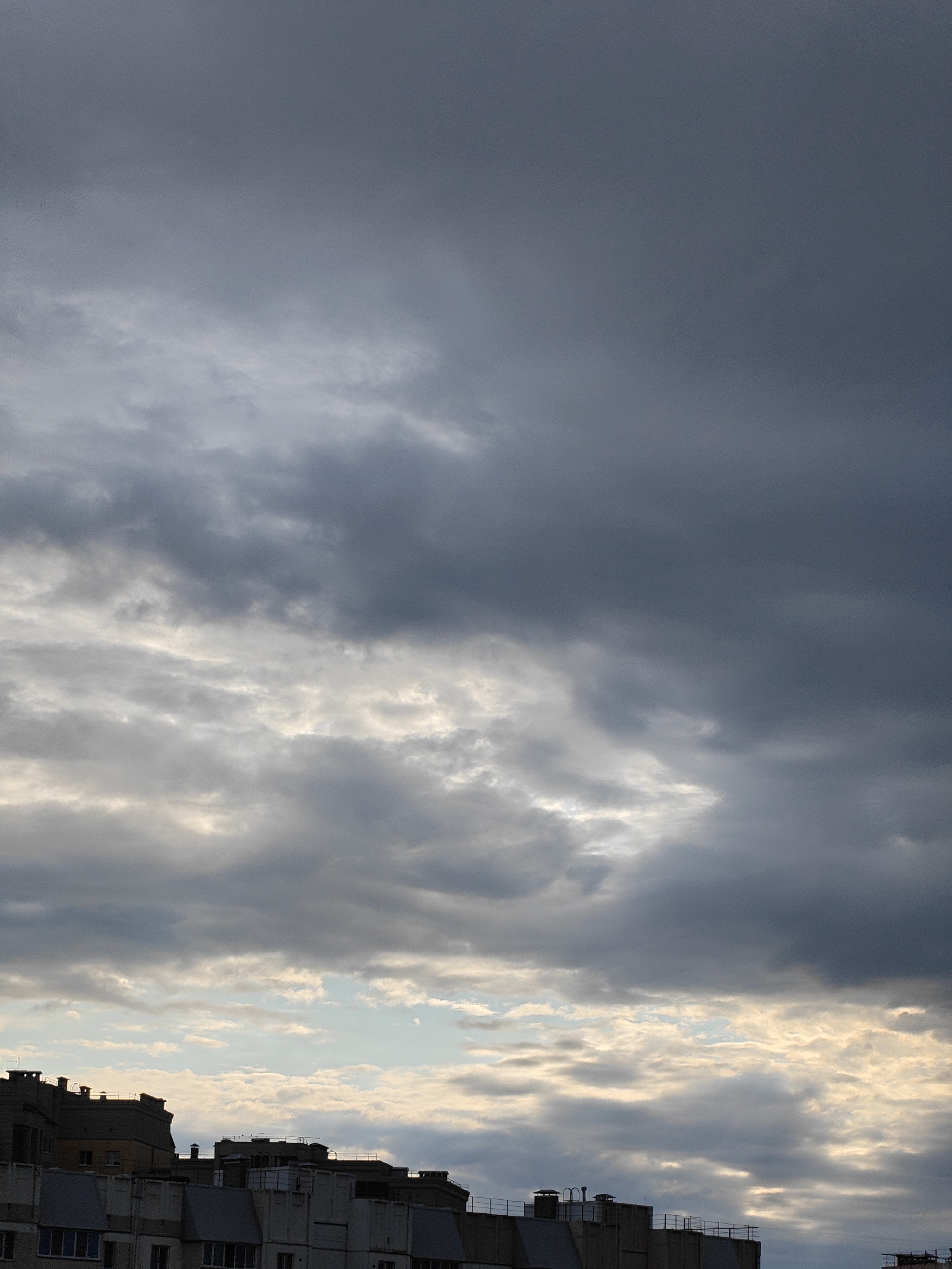 The image captures a cloudy sky above a row of apartment buildings in Krasnoobsk, Russia. The sky is filled with dark, heavy clouds, with some lighter patches indicating breaks in the cloud cover. The tops of several buildings are visible, featuring various architectural styles and rooftop details such as antennas and chimneys. The scene appears to be set during the daytime, though the overcast conditions suggest a possibly stormy or overcast weather. There are no people or visible activities depicted in the image.