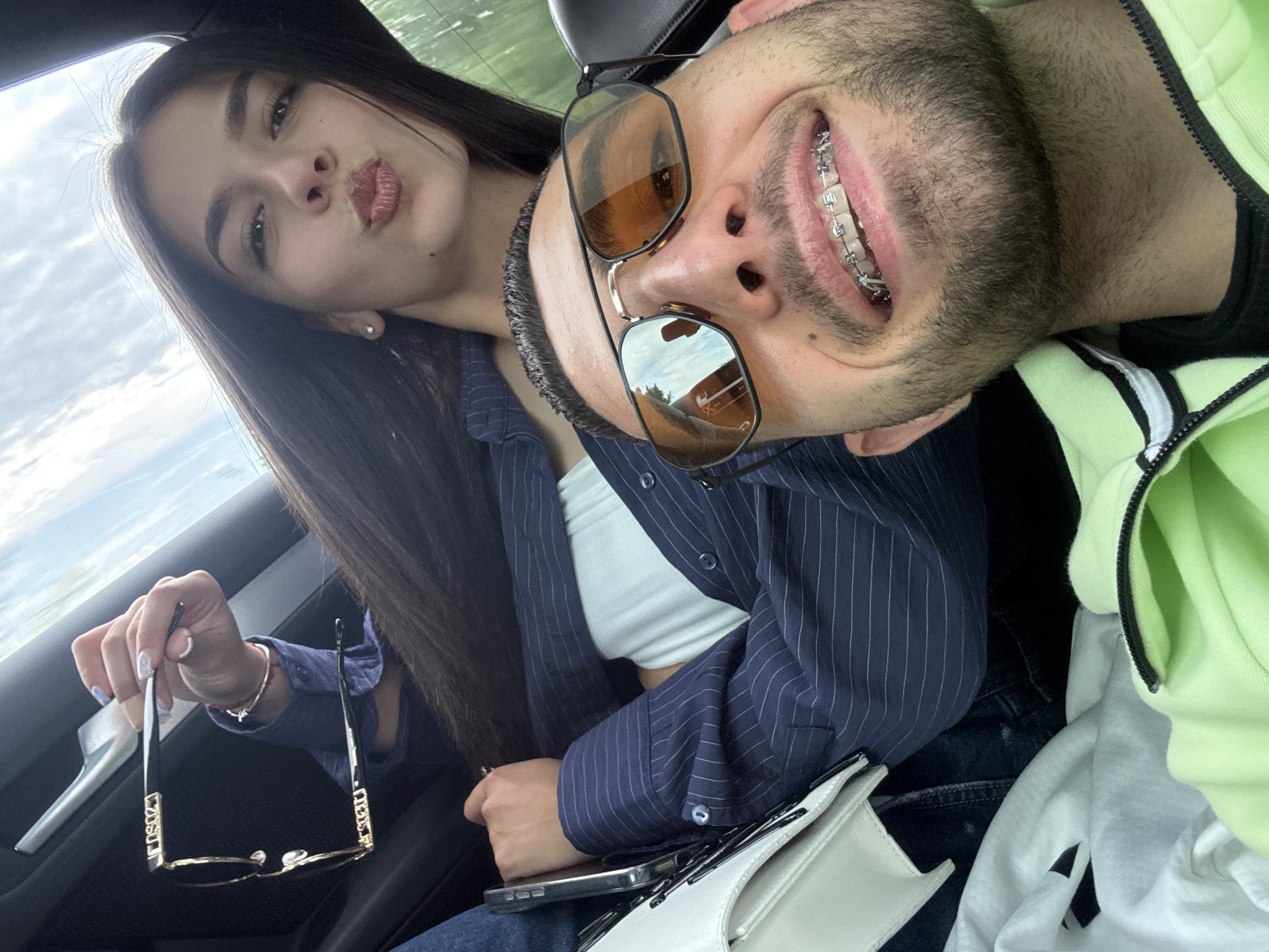 Two individuals are in the interior of a vehicle. The woman, with long dark hair, is wearing a blue striped shirt and is holding a pair of sunglasses in her hand. She is posing with pursed lips. The man has a beard and is wearing glasses on his head and a light green hoodie. He is smiling, revealing braces. The environment suggests a car interior, with parts of clothing, a white handbag, and a phone visible. The context of Polski Trambesh, Bulgaria, is not apparent within the visual information provided.