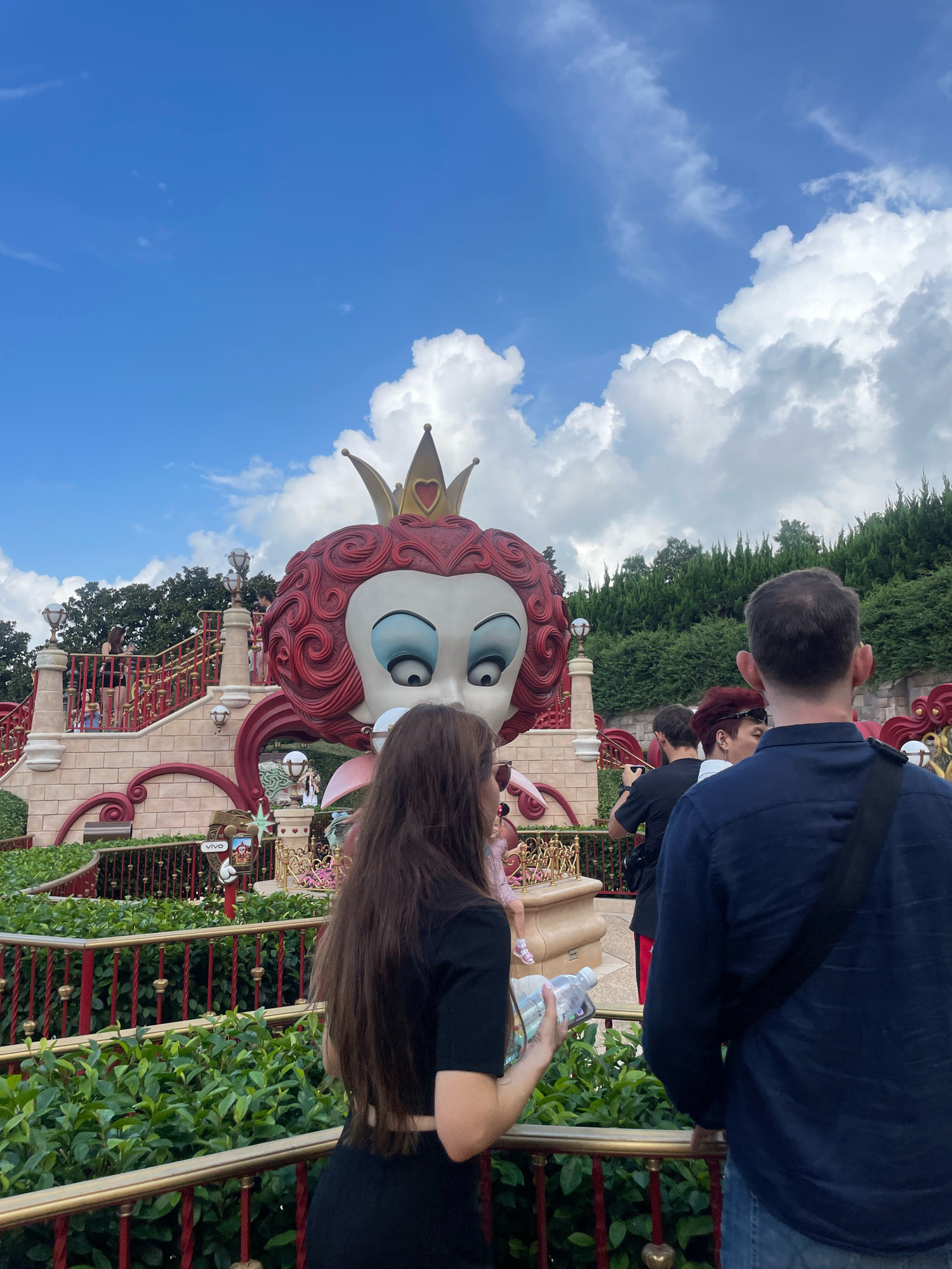 In Pudong, China, a large, stylized head of the Queen of Hearts from Alice in Wonderland dominates the scene, wearing a golden crown with a red heart emblem. The figure is part of a colorful, theatrical outdoor attraction. In the foreground, a woman with long brown hair, wearing a black top and skirt, holds a plastic water bottle and a smartphone. Behind her, a man in a dark blue button-down shirt and jeans stands facing away from the camera, with a black strap across his shoulder. Other individuals are visible in the background, on a red and gold tiered structure behind the Queen of Hearts. The environment includes lush green bushes and trees, with a bright blue sky dotted with white clouds above. The scene appears to be an amusement park or theme park setting.