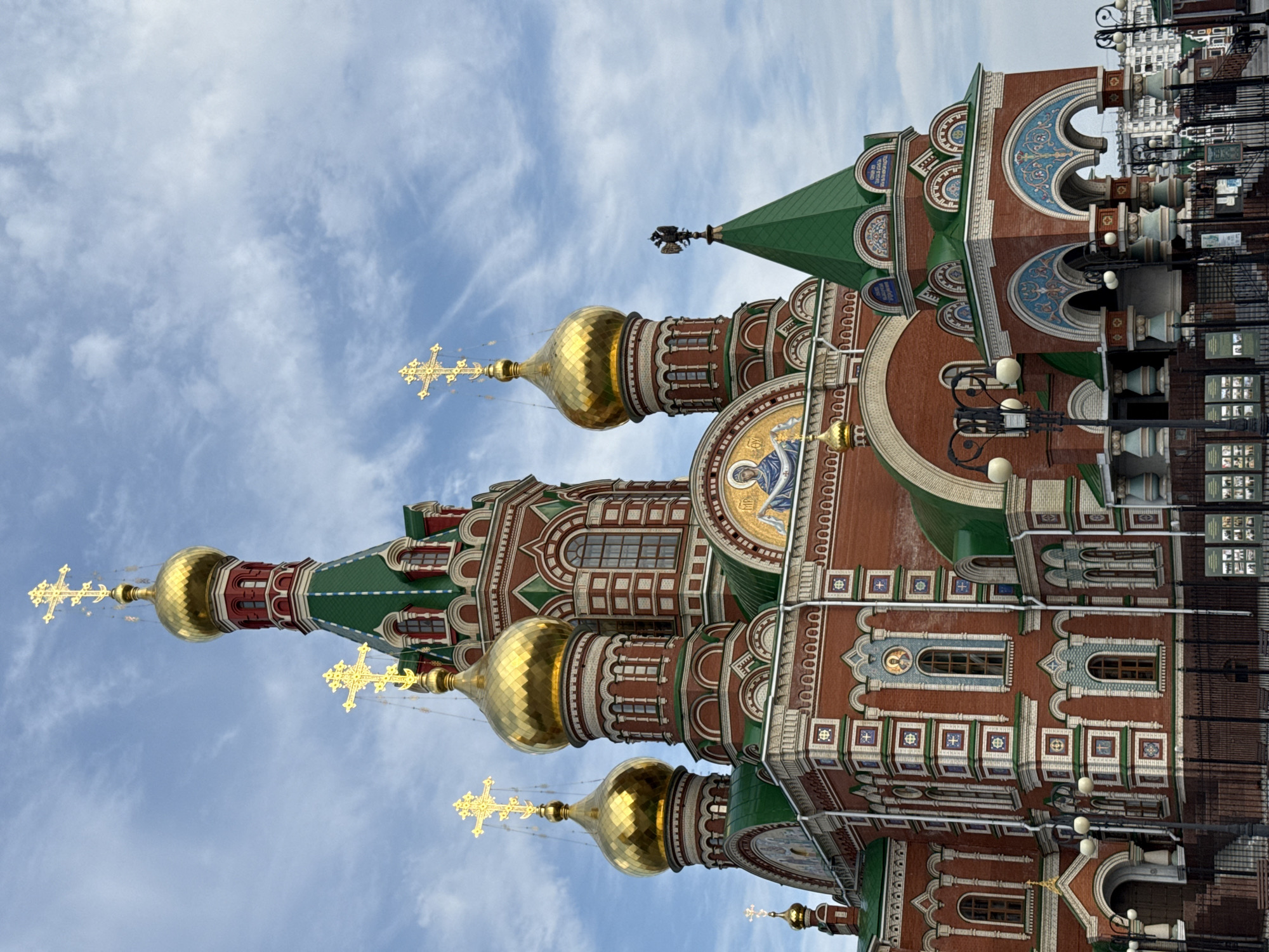A low-angle, close-up shot captures the brick exterior of the Resurrection Cathedral in Yoshkar-Ola, Russia. The cathedral features multiple golden domes topped with intricate crosses, and green conical roofs with smaller golden crosses. The red brick facade is adorned with decorative white and green brickwork, colorful tile mosaics depicting religious figures, and arched windows. The sky above is partly cloudy with streaks of white clouds. In the foreground, a black metal fence encloses the cathedral grounds, with lampposts visible behind it. Residential buildings and a cloudy sky are visible in the background. There are no people visible in the image.