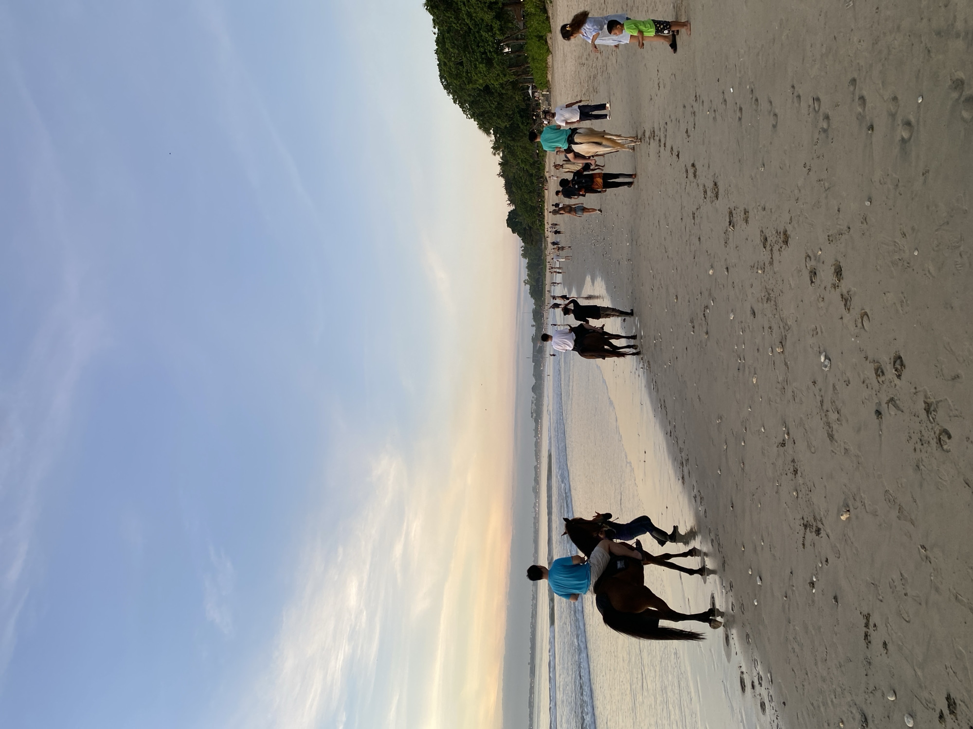 The image shows a beach scene, likely taken in the late afternoon given the lighting. A group of people is present on the beach. At the lower right, a person in a blue shirt is riding a dark horse along the water's edge. Further down the beach, groups of people are walking and standing near the shoreline. The environment appears to be a sandy beach with vegetation along one edge. Based on the location of Kuta Selatan, Indonesia, this is likely a beach in that area.