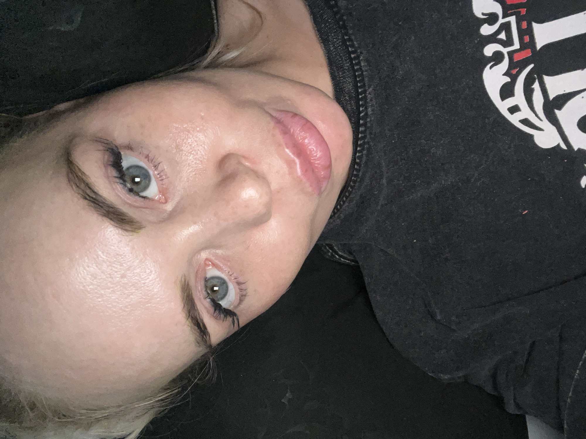 A close-up, overhead shot shows a woman's face from the nose up. She has light skin with some freckles and appears to be wearing makeup, including false eyelashes. Her eyes are light blue and her lips are slightly parted. Her hair is blonde and appears to be styled up. She is wearing a black t-shirt with a white and red design on it. The background is dark and appears to be a dark fabric.