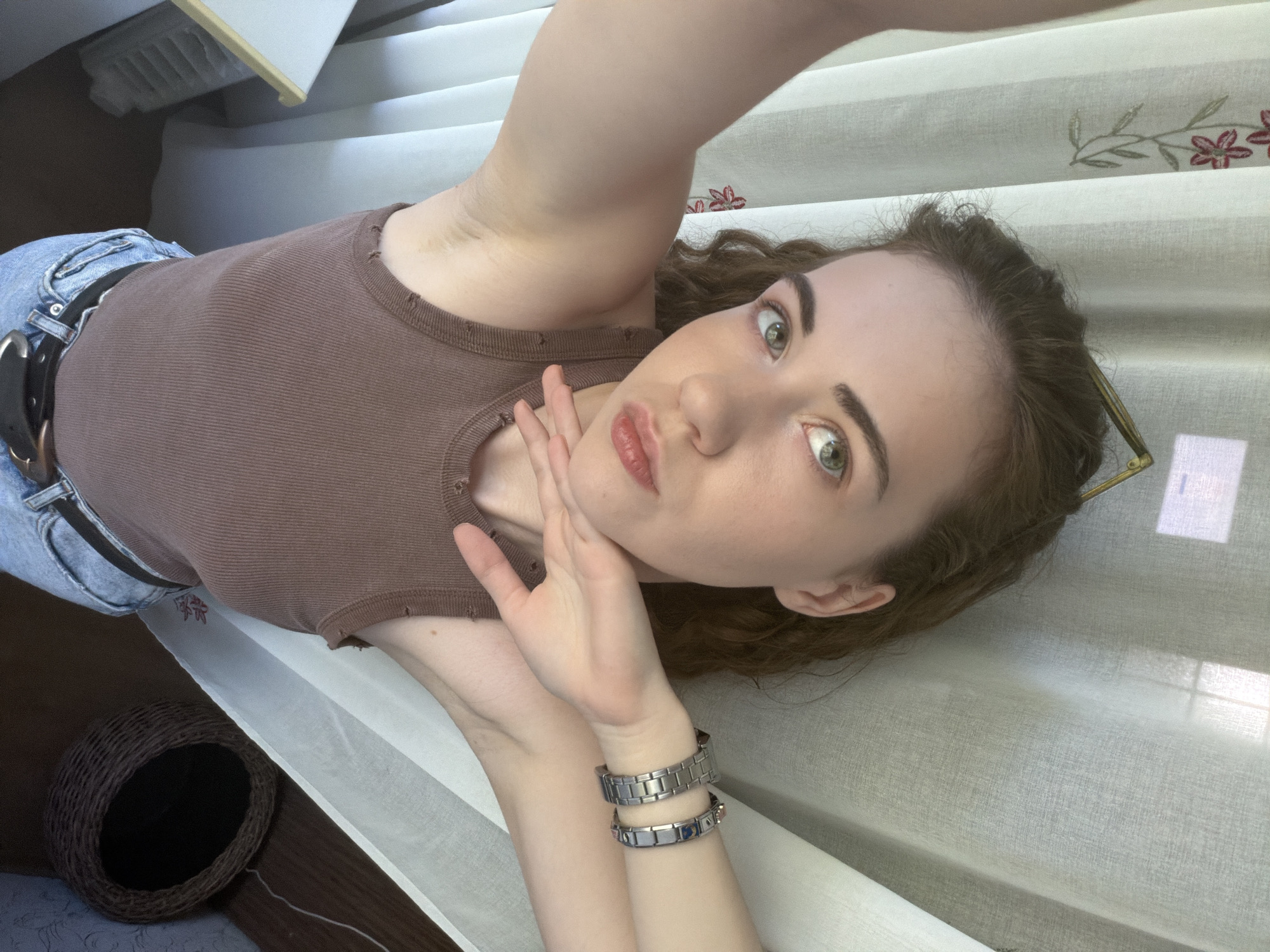 A young woman with curly brown hair and green eyes is taking a selfie. She is wearing a brown ribbed tank top and blue jeans with a black belt. She has on two bracelets on her left wrist. Her right arm is raised, and her hand is resting on her chin. She is looking at the camera with a pouting expression. The background appears to be indoors, with white curtains with a floral pattern. The lighting suggests it is daytime. There is no visible indication of the specific location within the city of Zavodoukovsk, Russia.