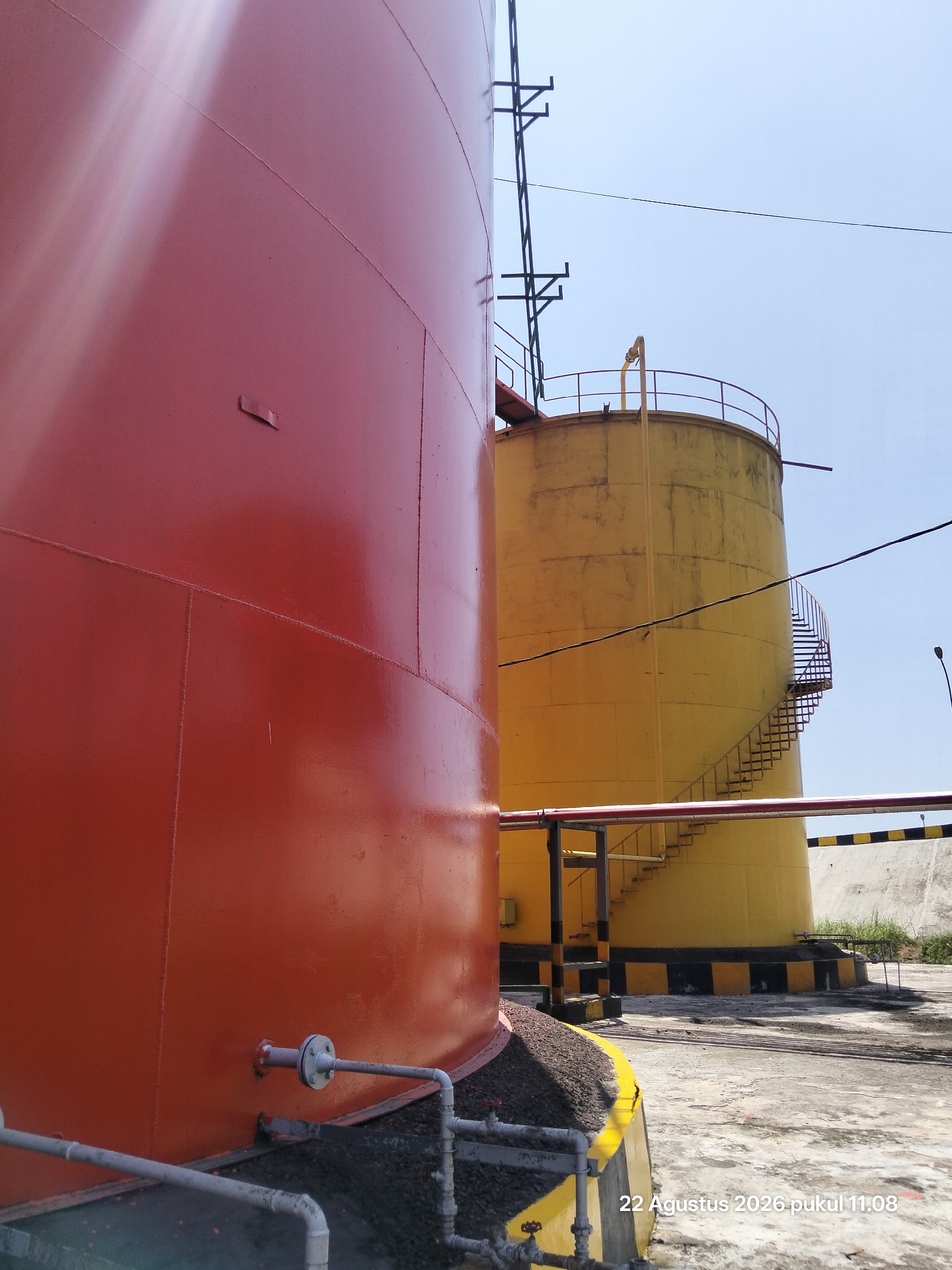 The image shows two industrial storage tanks outdoors. The tank in the foreground is large and painted bright red, with visible seams and a small metal protrusion on its side. A network of pipes and valves is visible near its base, running across the ground. In the background, a second, taller tank is painted yellow and shows signs of weathering. It has a spiral staircase leading up to a platform at its top, and a yellow pipe extends from the top. Both tanks are situated on a paved surface, with areas of dark gravel and some vegetation visible. A concrete barrier with black and yellow stripes runs along the edge of the yellow tank's base. The sky is clear blue. A timestamp "22 Agustus 2026 pukul 11.08" is visible in the bottom right corner. The scene appears to be an industrial facility, possibly a petroleum or chemical storage site.