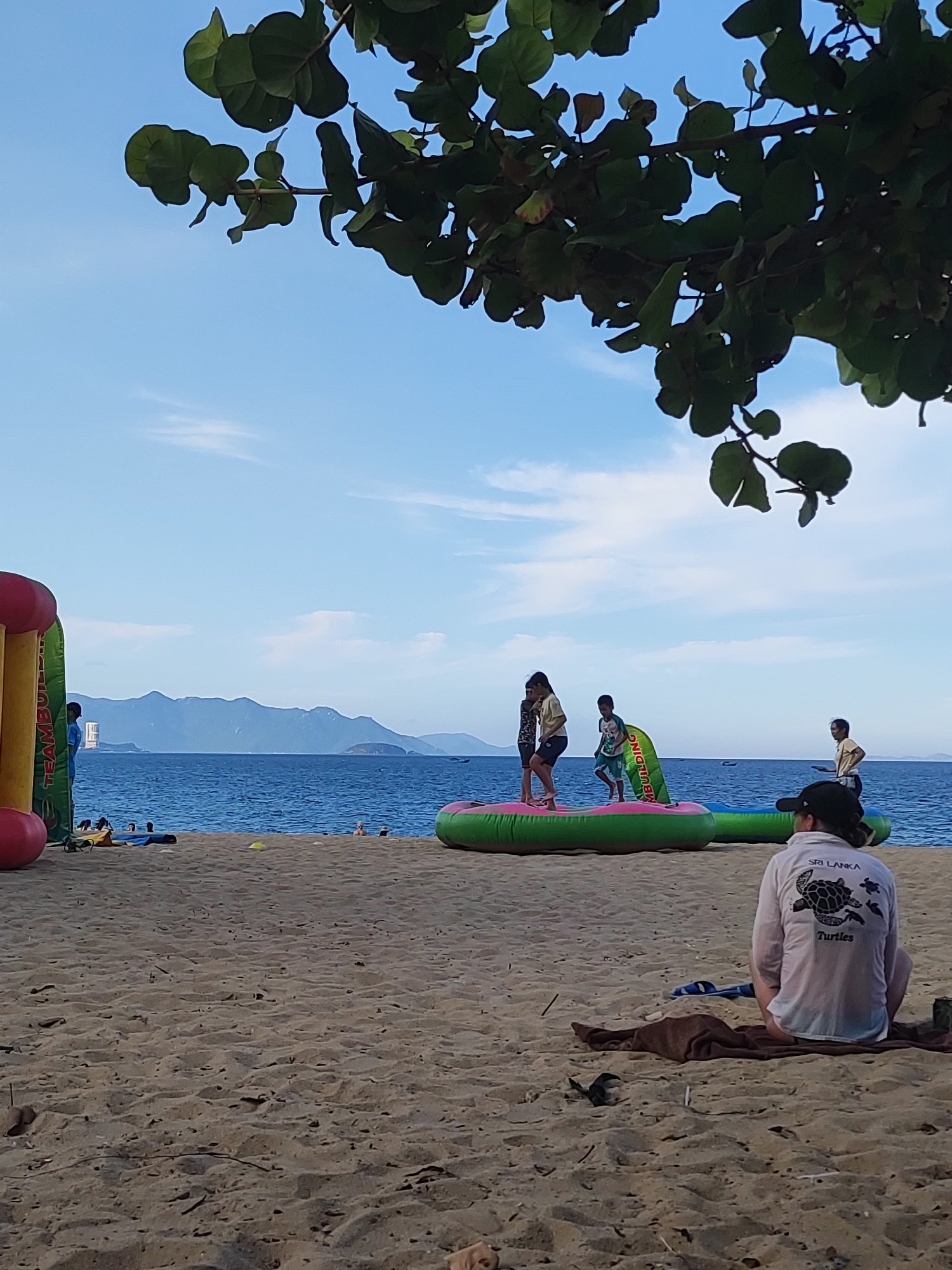 The image depicts a sunny day at a beach in Nha Trang Ward, Vietnam. In the foreground, a person is sitting on a towel on the sandy beach, facing away from the camera, wearing a light-colored shirt with a turtle graphic and the words "SRI LANKA Turtles." To the left, a colorful inflatable structure, partially visible, appears to be part of a water park attraction. Further back, several people are visible near the water's edge. Some are standing on another inflatable structure in the sea, possibly playing or relaxing. Children are among them. The sea is a vibrant blue, stretching towards a distant coastline with mountains. The sky is a clear, bright blue with a few wispy clouds. Green foliage from a tree overhangs the top of the image, framing the scene. The overall atmosphere suggests a recreational day at the beach.