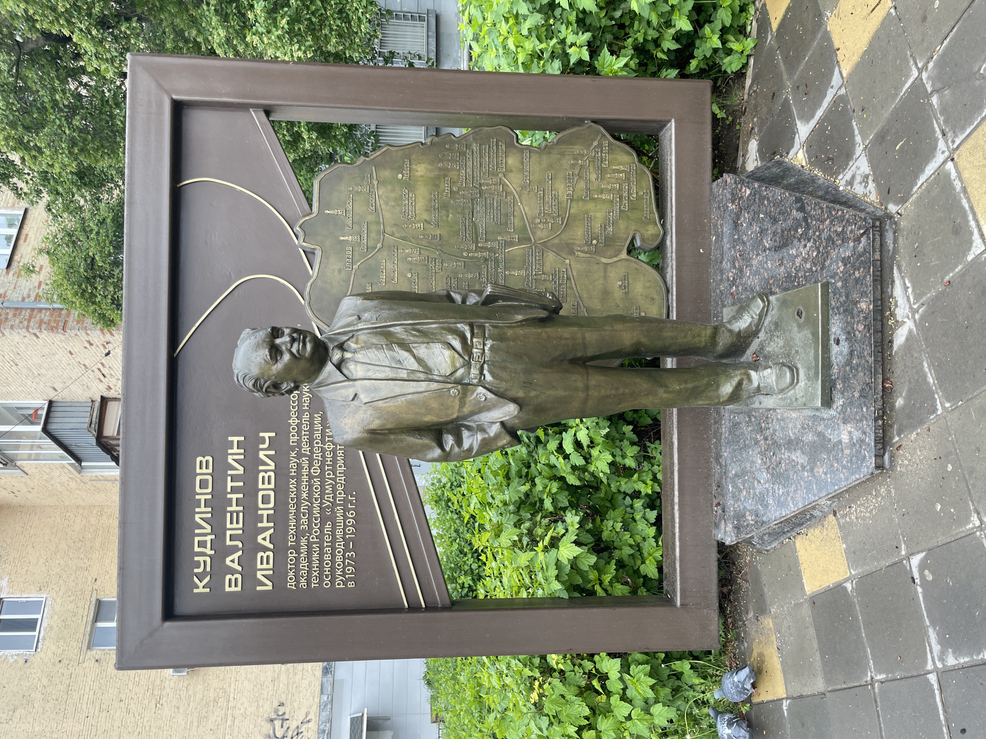 The image shows a bronze monument to Kudinov Valentin Ivanovich, a doctor of technical sciences, professor, academician, and founder of "Udmurtneft." The monument includes a bronze statue of the man, a map of Izhevsk, Russia, behind the statue, and a plaque with text in Russian. The monument is placed on a granite base surrounded by greenery and is located on a sidewalk paved with gray and yellow tiles.