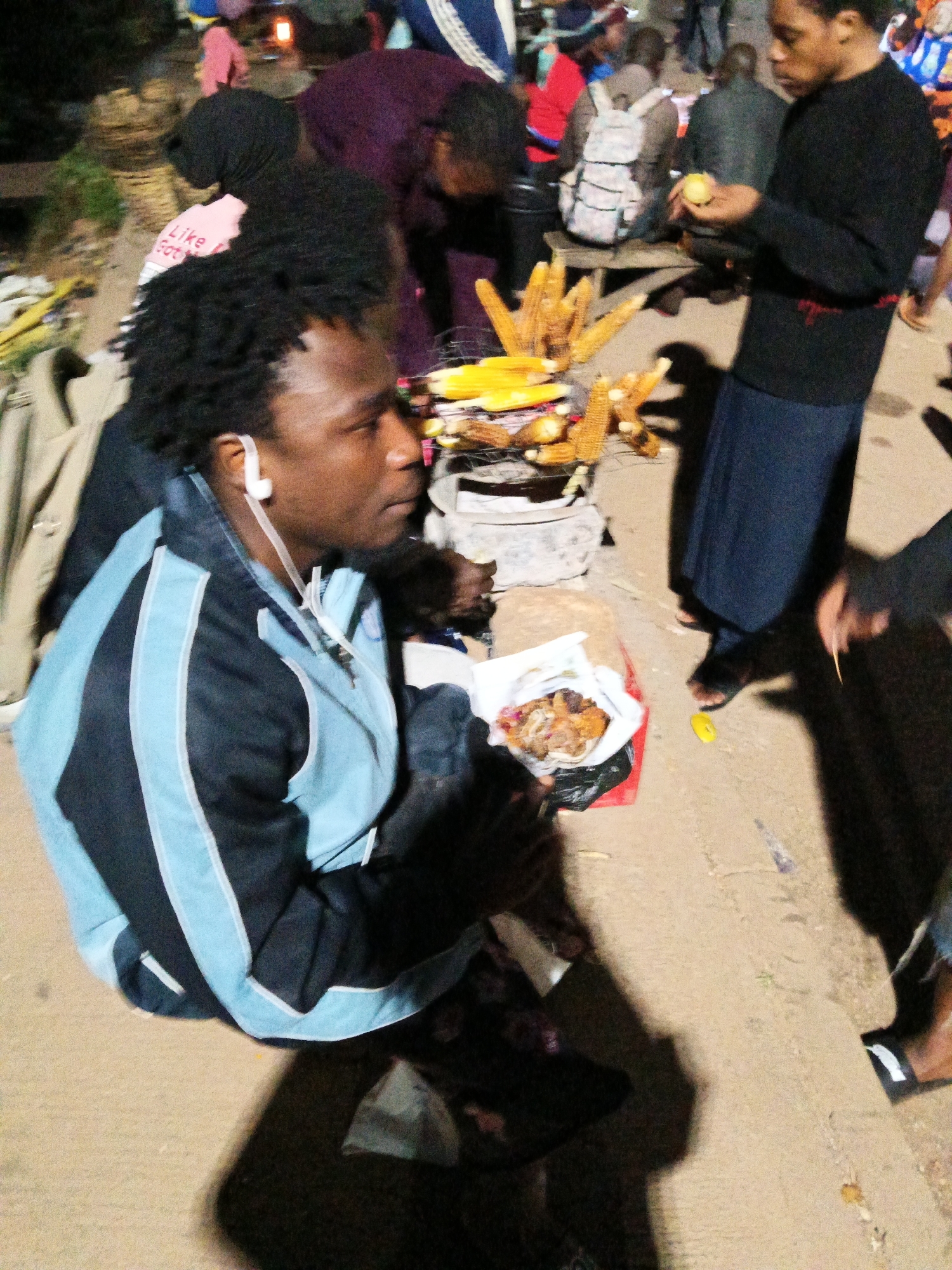In Bukuru, Nigeria, a street vendor is captured in a nighttime scene, the image focused on their upper body and the surrounding marketplace. The vendor, a man with dark, braided hair and a light-skinned complexion, is wearing a blue and black athletic jacket and appears to be listening to music through white earbuds. His expression is neutral as he sits, facing right, his hands occupied with items on a table or cart.
In the background, other individuals are visible. A person wearing a black hijab and a pink shirt with "Like God" printed on it is partially obscured. Further back, several people are milling about, some wearing backpacks, suggesting a busy market environment. A grill with several ears of corn on it is positioned to the right of the vendor, indicating food is being prepared or sold. Bags of food and other items are scattered around the scene. The ground is a mixture of sand and dirt, typical of an outdoor market. The lighting suggests it's evening or night, with artificial lights casting shadows.
