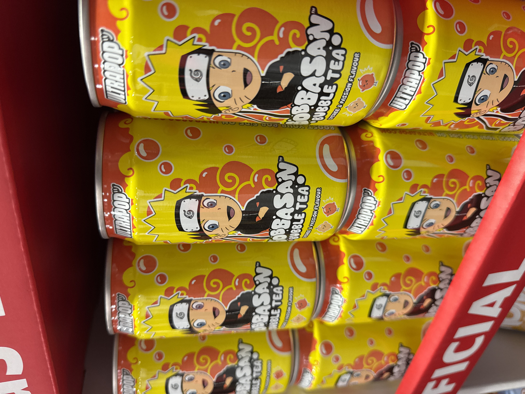 A vertical, eye-level, close-up shot showcases a shelf of vibrant yellow soda cans featuring cartoon characters in a retail environment. The cans, branded with "ULTRAPOP" and "BOBBASAN BUBBLE TEA" with "LYCHEE & PASSION FLAVOUR" text, are stacked and arranged on a red shelf. The character depicted on the cans appears to be a stylized anime character with spiky yellow hair, a black outfit, and a forehead protector. The background includes a blurred red shelf at the top and a red signage with white text reading "FFICIAL" at the bottom. The lighting is bright, suggesting an indoor setting like a supermarket or convenience store. The image does not depict any people, interactions, or specific environmental features of Sofia, Bulgaria.