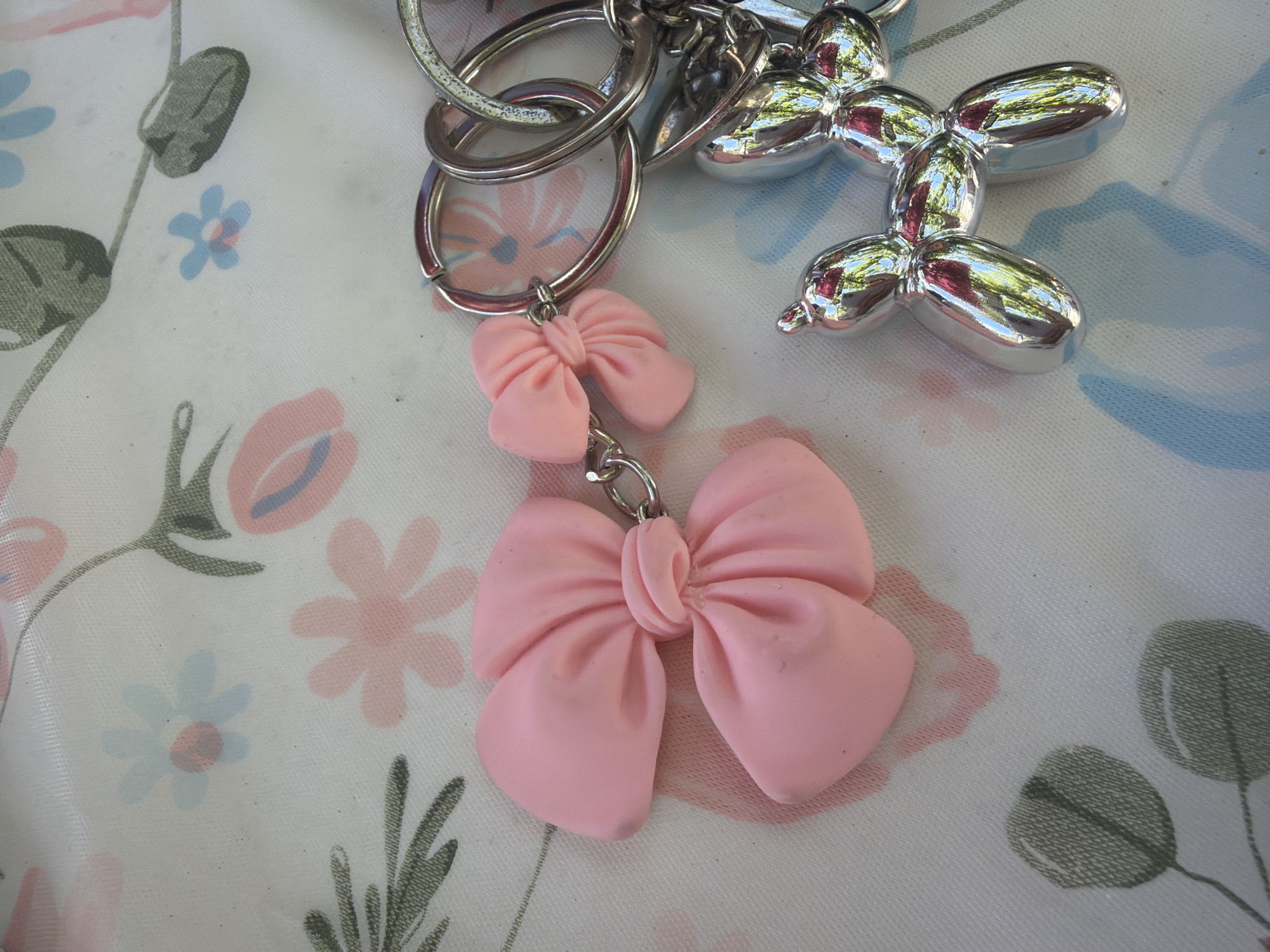 The image shows a close-up of a floral-patterned tablecloth, with a pink bow-shaped keychain and a silver balloon dog keychain as the main subjects. The tablecloth has a white background with scattered stylized flowers in pink and blue, and green leaves and stems. The pink bow keychain is large and appears to be made of clay or a similar material, with a smaller pink bow attached to it via a silver chain and ring. The silver balloon dog keychain is highly reflective, showing distorted reflections of the surroundings, which suggest it is made of polished metal. It is also attached to silver keyrings. The lighting suggests the image was taken outdoors, possibly in sunlight, as indicated by the shadows cast by the keychains. There is no indication of the city of Vlasatitsa, Bulgaria, in the image itself.
