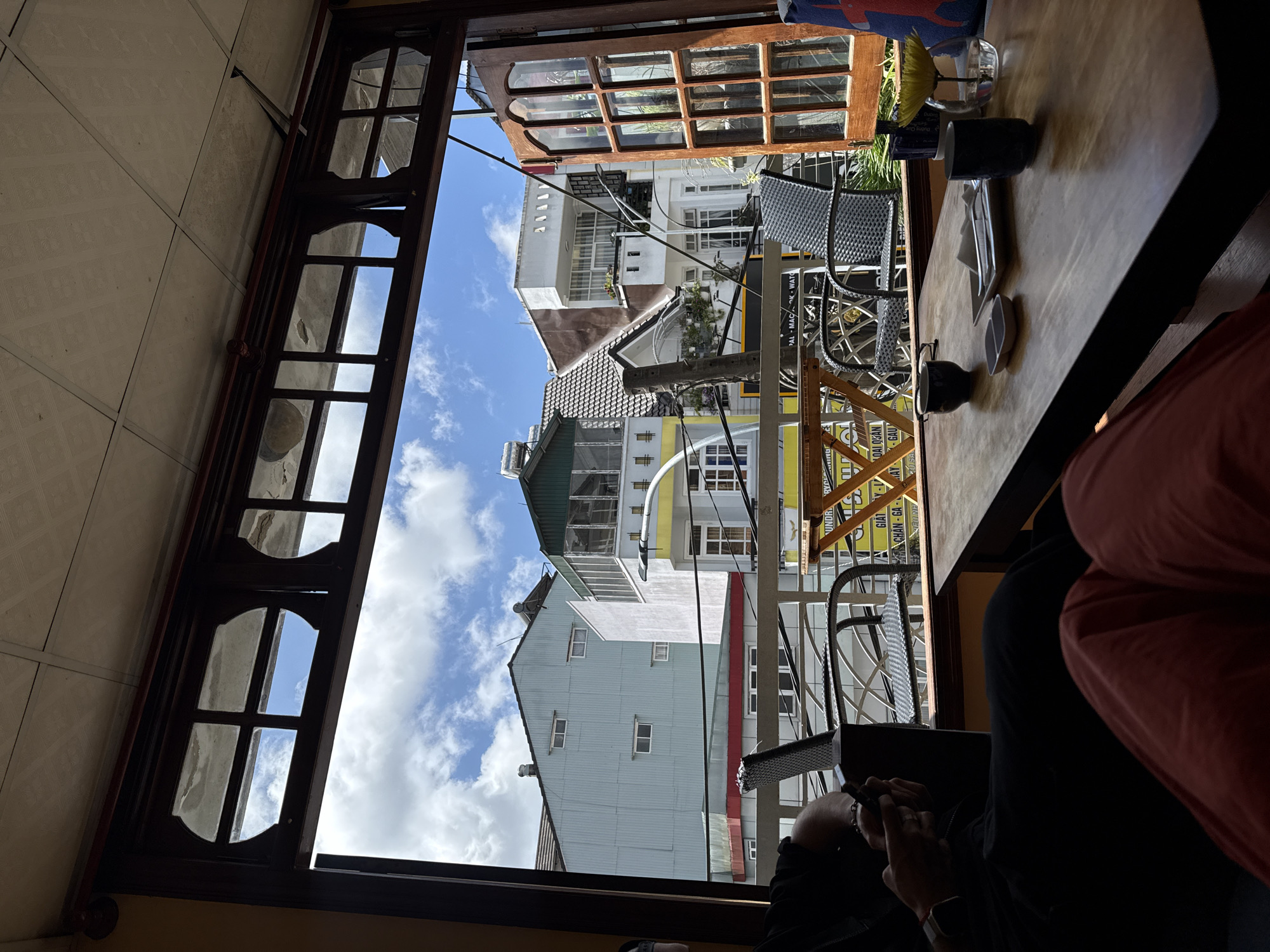 This image depicts an indoor cafe scene with a view of Da Lat, Vietnam. The perspective is from a seated person looking out a large, multi-paned window. The window frame is dark wood with decorative circular and rectangular panes. Through the window, a street scene with several buildings is visible under a bright, cloudy sky. The buildings appear to be of varying architectural styles and colors, with some having balconies and windows. There is a wooden table in the foreground with two black mugs, a small dish, and a plate with cutlery. A person's lap, covered in red fabric, is in the lower part of the frame, indicating the viewer's position. The upper part of the window frame shows a textured ceiling. Outside, a balcony area with chairs and tables is visible, along with a wooden door on the right side of the frame. The scene suggests a relaxed atmosphere within a cafe.