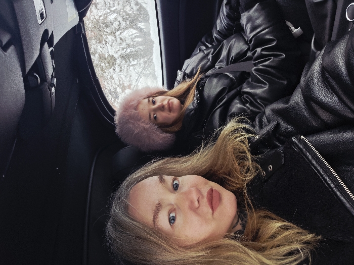 Two women are inside a car. One woman with blonde hair is positioned in the foreground, wearing a black leather jacket and a hint of a neck tattoo is visible. Behind her, a younger woman wears a pink fur hat and a black puffer jacket with a seatbelt across her chest. Outside the car window, a snow-covered landscape is visible. The image appears to be taken at an angle.