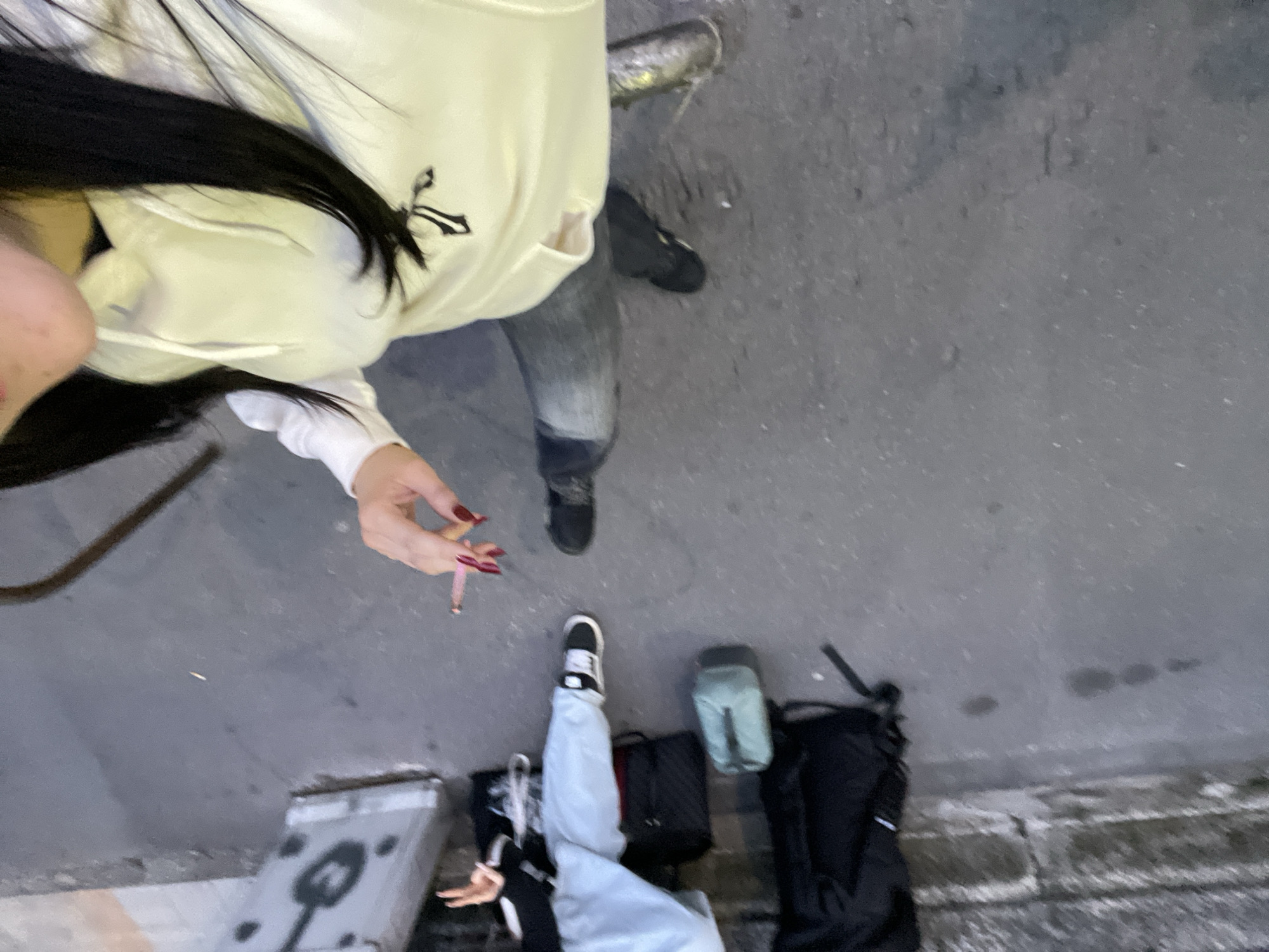 The image, taken from an overhead perspective, shows a partial view of two individuals and their belongings on a gray concrete or asphalt surface, likely a sidewalk in Milan, Italy. One person is wearing a light-colored hoodie, faded jeans, and black shoes, holding a lit cigarette in their hand. The other person is wearing light blue pants and black-and-white shoes. Near the second person are two black bags, a light green pouch, and a silver metal box or container. The background consists of the gray surface and what appears to be a textured wall along the edge of the frame. The image suggests an urban setting, possibly in Milan, Italy, given the likely context of the prompt.
