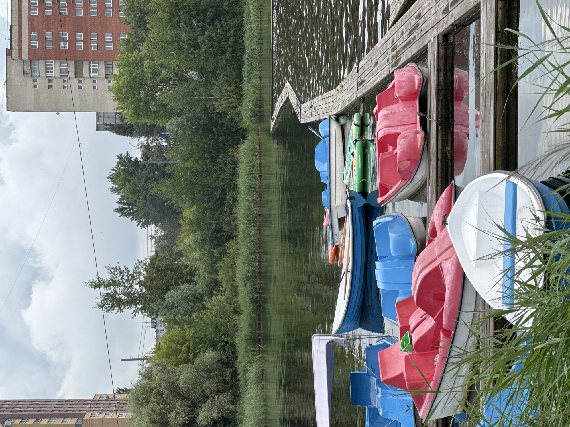 The image shows a collection of colorful paddle boats and a small rowboat docked on a wooden pier on a lake. The boats are of various colors, including red, blue, and white. Some of the boats are pedal boats, while one appears to be a rowboat with oars. The pier is made of weathered wooden planks and extends into the water. In the background, there is a dense line of trees and reeds along the edge of the lake. Beyond the trees, several multi-story buildings, some with brick facades and others with lighter colored exteriors, are visible, indicating an urban setting. The sky is overcast with visible clouds. This scene is consistent with a recreational area located in the city of Nizhny Novgorod, Russia, specifically near the Nizhny Novgorod Kremlin, which overlooks the confluence of the Volga and Oka rivers and has similar waterfront areas with recreational boat rentals. There are no people visible in the image.