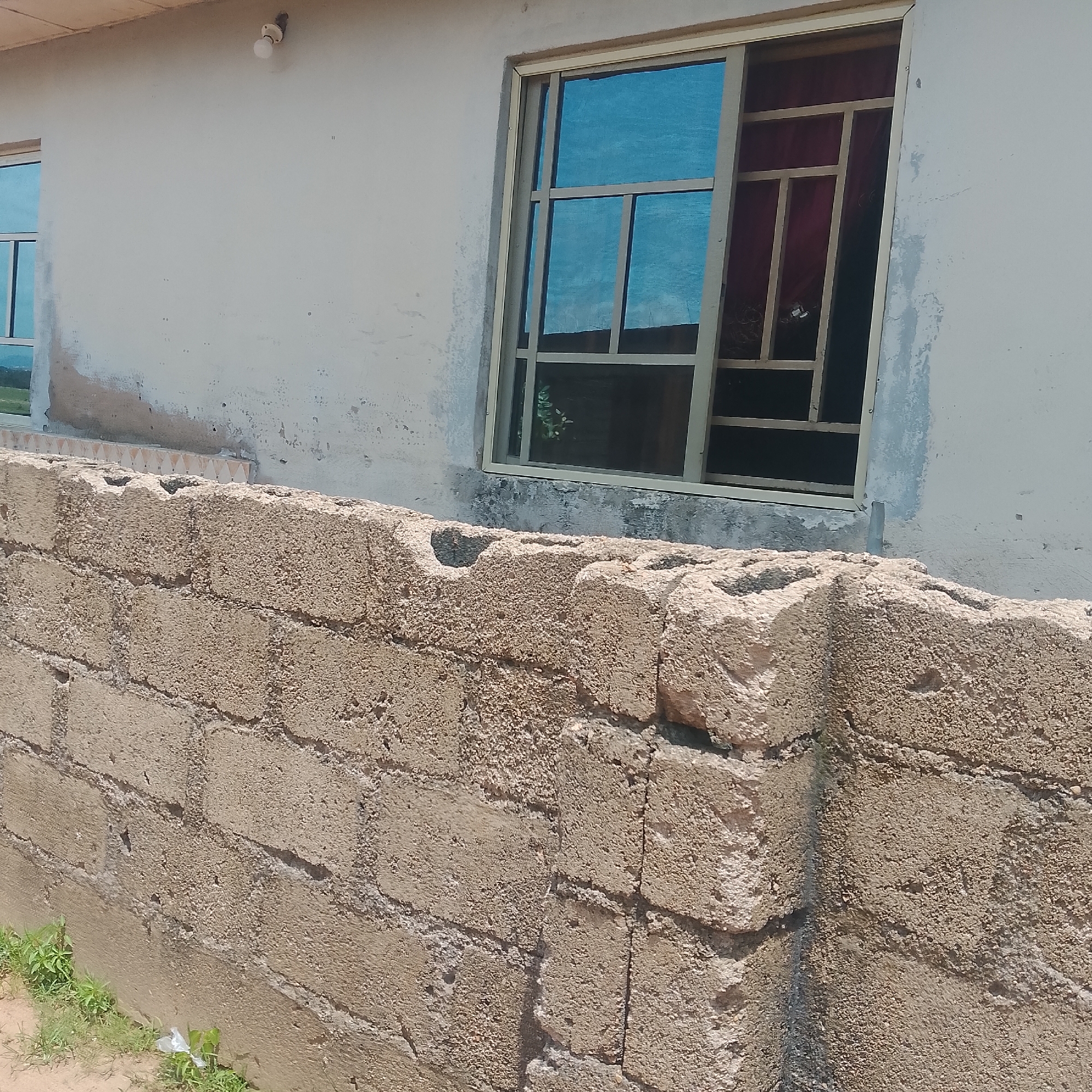 The image shows a portion of an unfinished building in Jalingo, Nigeria. In the foreground, a rough wall constructed from concrete blocks is visible. The blocks are unevenly textured and mortared together. Behind this wall, a portion of the building's exterior wall is visible, painted a light, off-white color. There are visible patches of dampness or discoloration on this wall. A large window with a metallic frame and blue-tinted glass is set into the building's wall. The window has a grid pattern inside the frame. To the left of this window, a portion of another window is visible. A single light bulb with a visible filament is screwed into a fixture on the exterior wall near the top edge of the frame. The ground in front of the block wall is sandy and sparse green vegetation is growing.