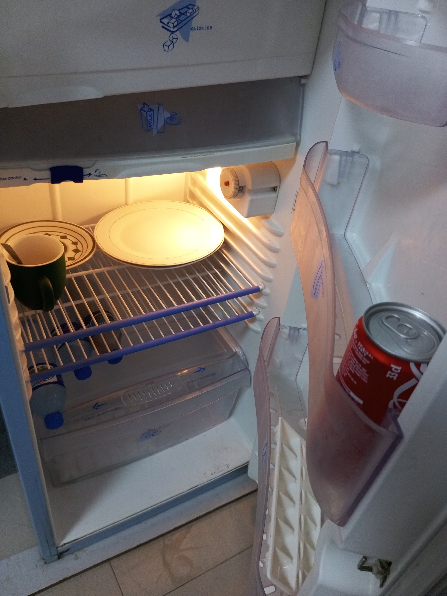 The image shows the interior of a refrigerator. On the top shelf, there is a green mug with a spoon in it, placed next to a plate. To the right of the plate is a large, white, round dish. The middle shelf has a clear plastic storage container with a blue lid. Below this container, there are two plastic bottles with blue caps. On the door shelf, a red can of Coca-Cola is visible. The refrigerator has a light that is turned on. The location of Bukuru, Nigeria, is not visually represented in the image.