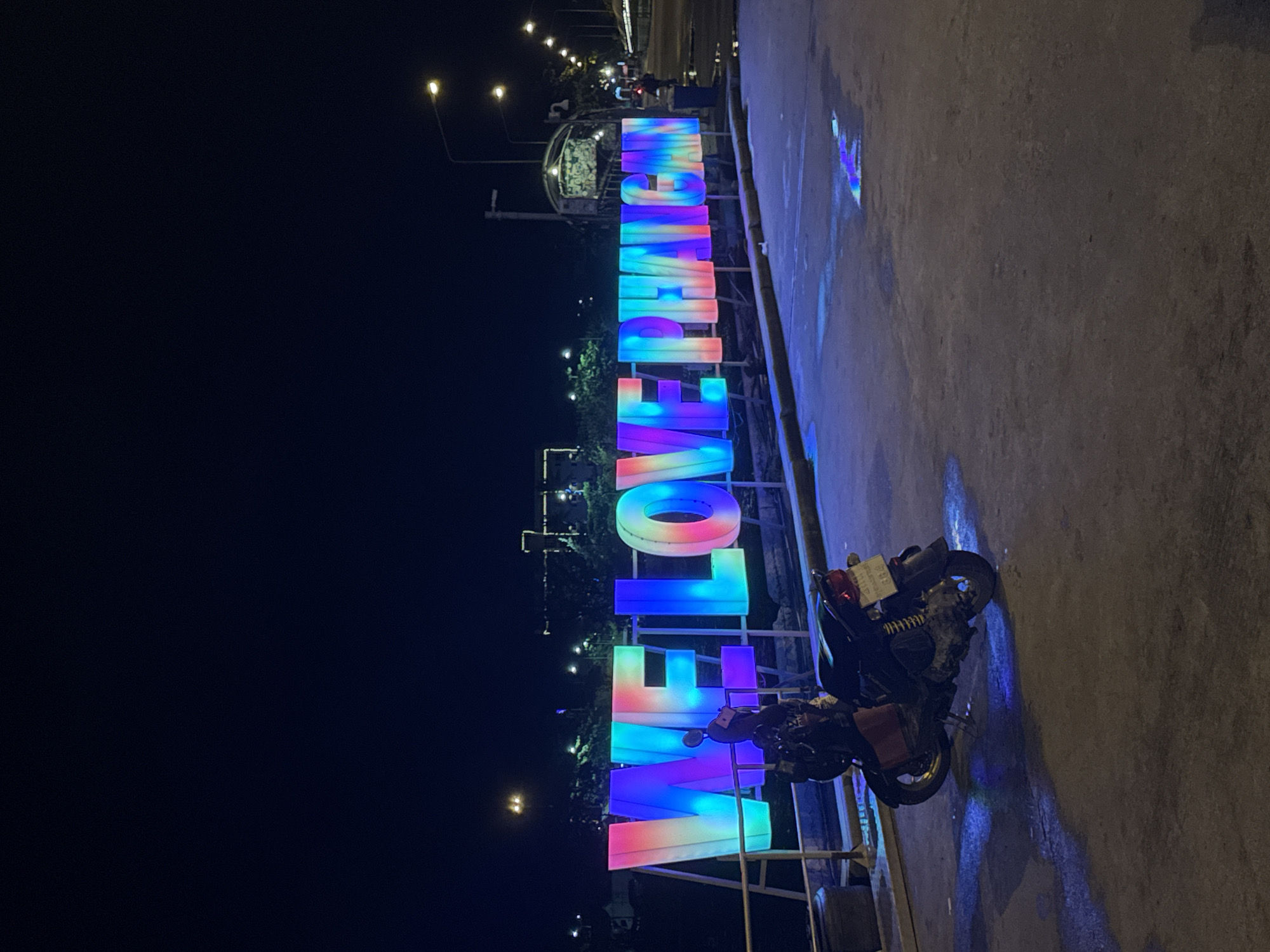 The image depicts a nighttime scene in ตำบลเกาะพะงัน, Thailand, featuring a large, illuminated sign spelling "WE LOVE PHA NGAN" in prominent capital letters. Each letter is internally lit with a vibrant, shifting gradient of colors including blue, purple, pink, orange, and green. The sign is positioned above a low railing or barrier, possibly overlooking a body of water not visible in the frame. In the immediate foreground, a dark-colored scooter is parked on a dark asphalt or concrete surface, facing away from the viewer. The scooter has a reddish-brown object on its seat area. The ground shows wet patches that reflect the colorful light from the sign, primarily in shades of blue and purple. In the background, across a dark expanse, distant buildings and structures are faintly visible with their own illumination. To the far right, a street extends into the distance, lined with multiple streetlights emitting a warm yellow glow. The sky is dark, indicating a nighttime setting. No people are explicitly visible, though the presence of the scooter and distant streetlights implies human activity.