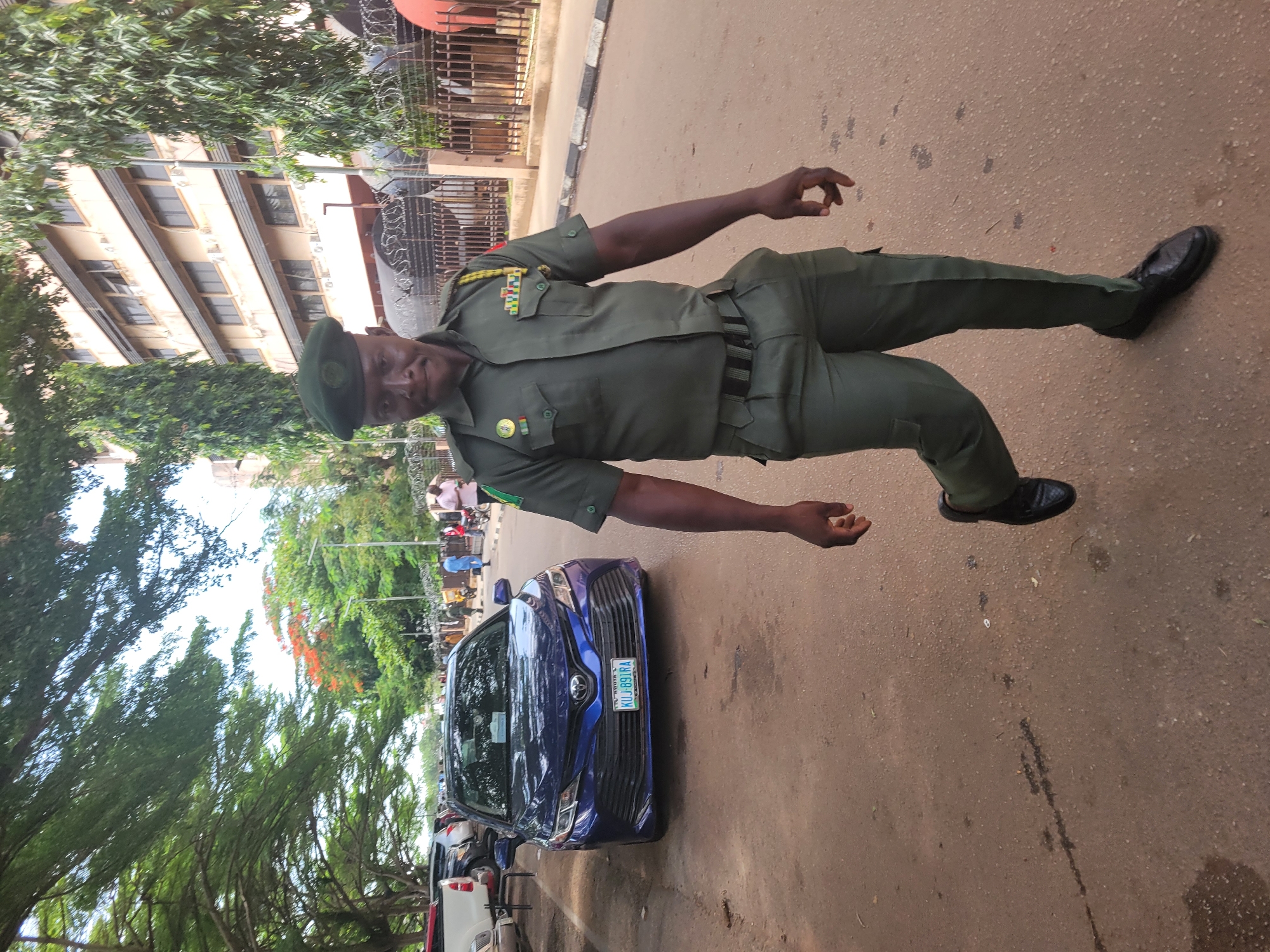 A man in an olive green military uniform stands on an asphalt road next to a blue Toyota Camry with a Nigerian license plate. The license plate reads "KUJ-891RA" with "Nigeria" printed above the number. Behind the car and along the curb, there are other parked vehicles, including a white one. There is a tree to the left and behind the man and car. In the background, a multistory building can be seen, partially obscured by trees and with what appears to be a barbed-wire fence along the top. People are walking in the background, further back from the man and the car. The location is in Abuja, Nigeria.