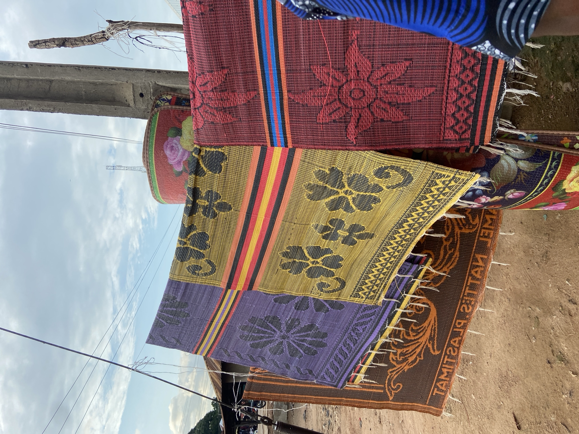 This image depicts an outdoor display of colorful, woven plastic mats for sale, likely in a market or roadside setting in Mararaba, Nigeria. Four distinct mats hang vertically, from left to right: a brown mat with an orange scroll pattern and the mirrored text "PLASTIC MATS"; a purple mat with a large black floral design and horizontal black, yellow, red, and purple stripes; a yellow mat featuring black four-petal floral patterns and horizontal black, yellow, red, and orange stripes; and a red mat with a red floral design and horizontal black, blue, orange, and red stripes. All hanging mats are fringed with white material at their bottom edges. A rolled-up mat, predominantly red with a large pink rose and green leaves, is positioned at the top center-right.
The mats are suspended from a wire or rope, which is supported by a dark metal pole on the left and a gray concrete utility pole on the right. White strings and frayed material are visible tied to the poles and wires. The ground beneath the mats is exposed reddish-brown earth, characteristic of unpaved areas. In the background on the left, a building with a corrugated metal roof and a partial view of a motorcycle are discernible. On the right, a slender, distant communication tower stands against the sky. The sky is light blue with scattered white clouds. The rightmost edge of the frame shows the right arm and torso of a person, clad in a blue garment with black stripes, suggesting human presence near the display. The lighting indicates daytime conditions.