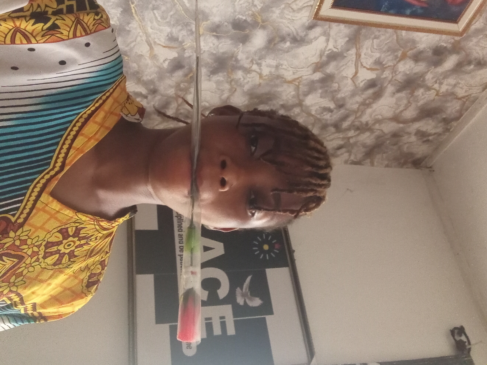 A person with braided hair is visible from the chest up, wearing a patterned garment with yellow, blue, and white sections. A clear plastic tube containing a red, green, and white substance is positioned vertically in front of the person's face, partially obscuring the view. Behind the person and the tube is a wall with a picture frame towards the top and a sign reading "ACE" with a sun-like symbol and a bird graphic. The location is Unknown, Unknown.