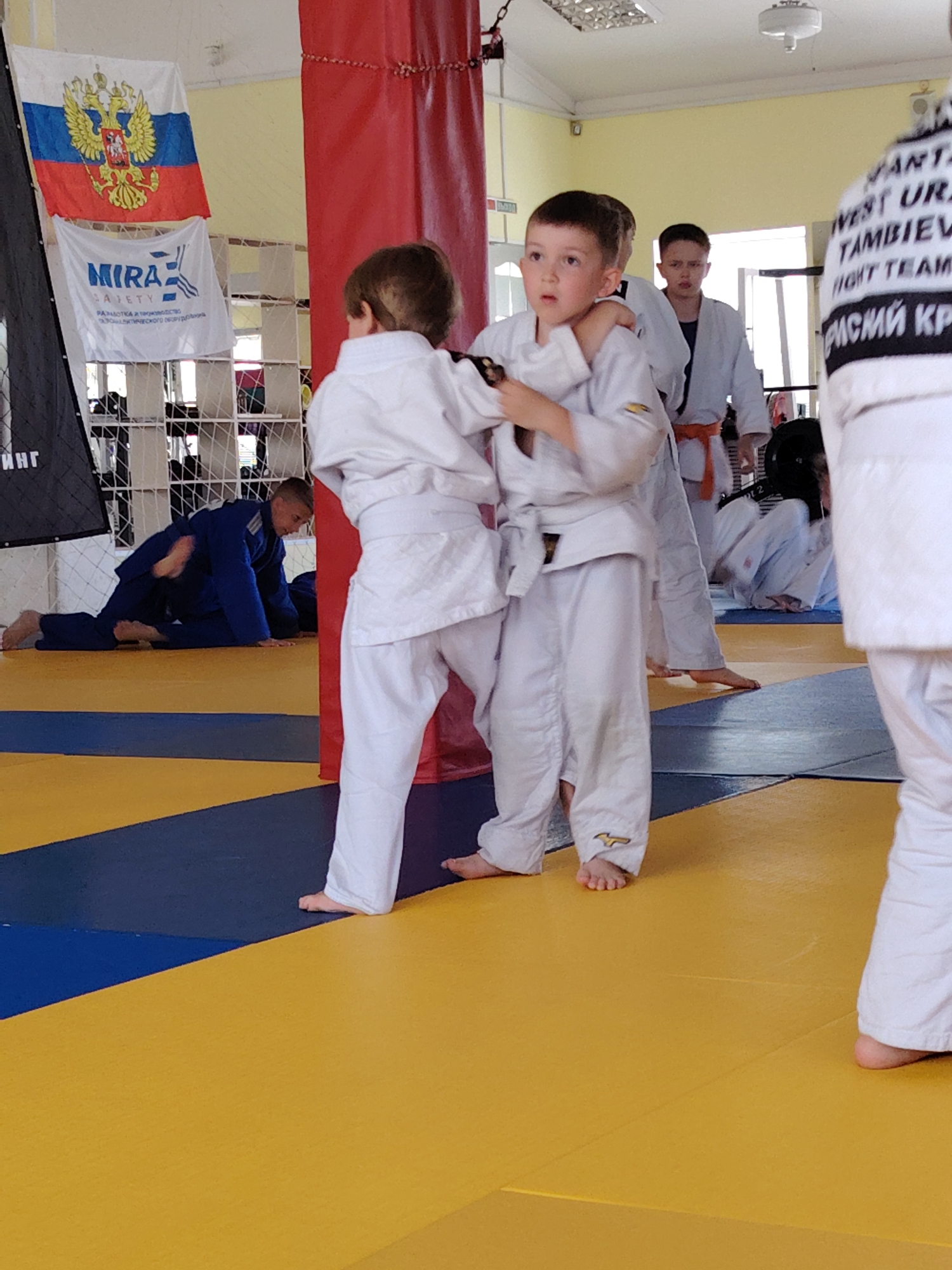 This image depicts an indoor martial arts training session, likely judo or sambo, taking place in Chaikovsky, Russia. The scene is set in a brightly lit gym or dojo with a floor covered in interlocking yellow and blue mats.
In the foreground, two young boys, both wearing traditional white martial arts gis (judogis/kimonos), are engaged in grappling practice. One boy has a firm grip on his partner's gi near the neck and shoulder, while the other is holding his opponent's sleeve. Their expressions are focused, indicating concentration on their technique.
In the mid-ground, other participants are visible. To the left, a person in a blue gi is on the mat, possibly practicing ground work or resting. Another child in a white gi with an orange belt stands observing. Further to the right, a person in a white gi wearing a black head protector is seated on the mat.
The background features a white wall with grid-like shelving. A red padded column, typical in martial arts training facilities, stands prominently in the center-right. Hanging on the left wall are two flags: the national flag of Russia (tricolor with the double-headed eagle emblem) and a white banner below it. This banner displays the logo and text "MIRA SAFETY" followed by Russian text that reads "РАЗРАБОТКА И ПРОИЗВОДСТВО" (Development and Production) and partially visible words related to "оборудования" (equipment). On the back of a white gi, partially seen on the far right, text includes "SPARTA," "WEST URAL," "TAMBIEV," "FIGHT TEAM," and "КРИСИЙ КР." The atmosphere suggests a daytime training session focused on youth development in martial arts.