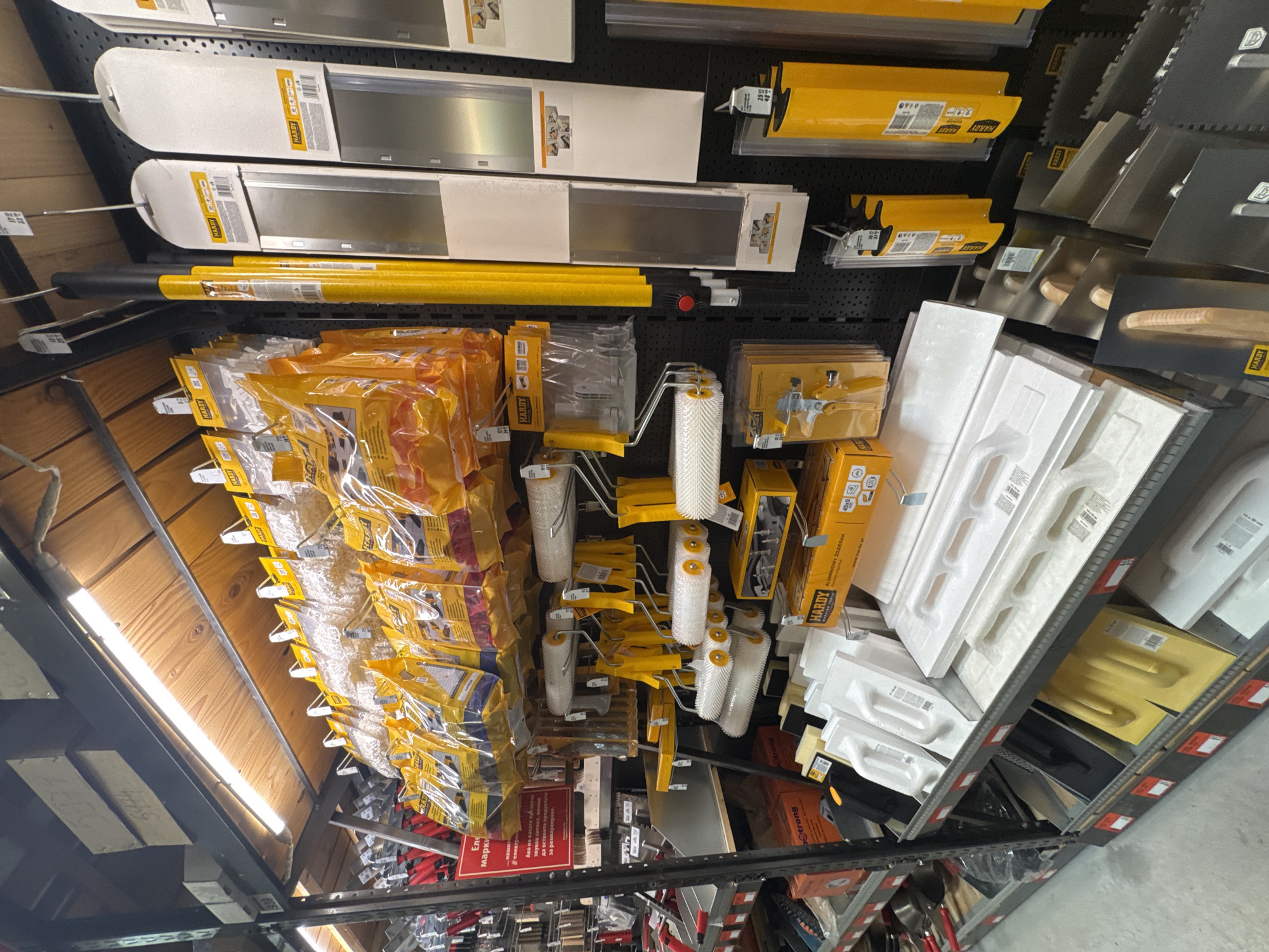 This appears to be a view of a shelving unit in a store, most likely a hardware or home improvement store. The shelves are stocked with various tools and supplies, with a focus on items related to painting, plastering, and wall finishing. Some of the items visible include:
* Various lengths of wall scrapers/blades, some packaged in white boxes.
* Packages of paint rollers, some still in their plastic wrapping.
* Plastering trowels, some with wooden handles.
* Extension poles, possibly for paint rollers.
* Packages and boxes labelled "HARDY," which appears to be a brand name.
* An information sign in Bulgarian, including a warning about an alarm sounding.
The overall impression is of a well-stocked section of a store catering to DIY enthusiasts or professional tradespeople. The shelves themselves are metal with a perforated back panel. The viewpoint is tilted upwards, giving a somewhat distorted perspective. The location is inferred from the sign to be in Bulgaria, presumably in the city of Sofia.